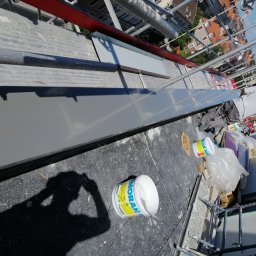 Paweł Zaborowski - Praca na wysokości: blacharz na rusztowaniu przy montażu szarej obróbki blacharskiej na dachu, widoczne puszki z farbą i narzędzia, cień fotografa w lewym dolnym rogu.