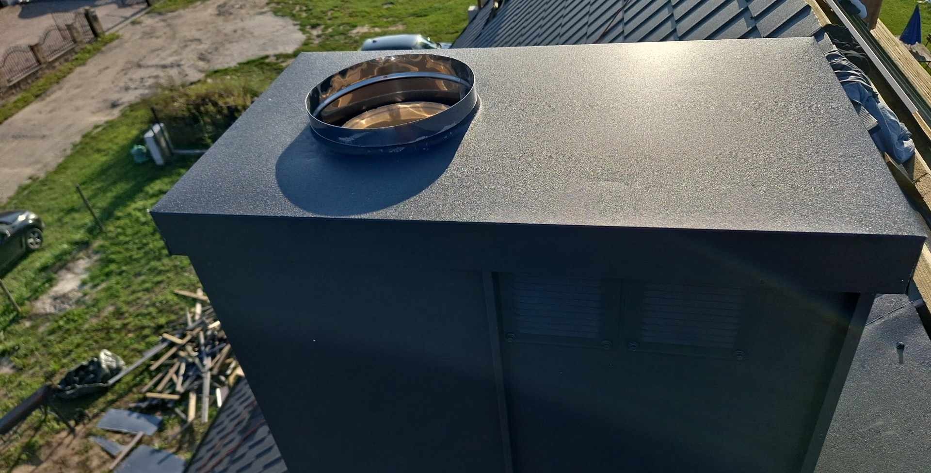 Nowoczesny, czarny komin z metalowym wylotem na tle dachu i zieleni, widok z góry.