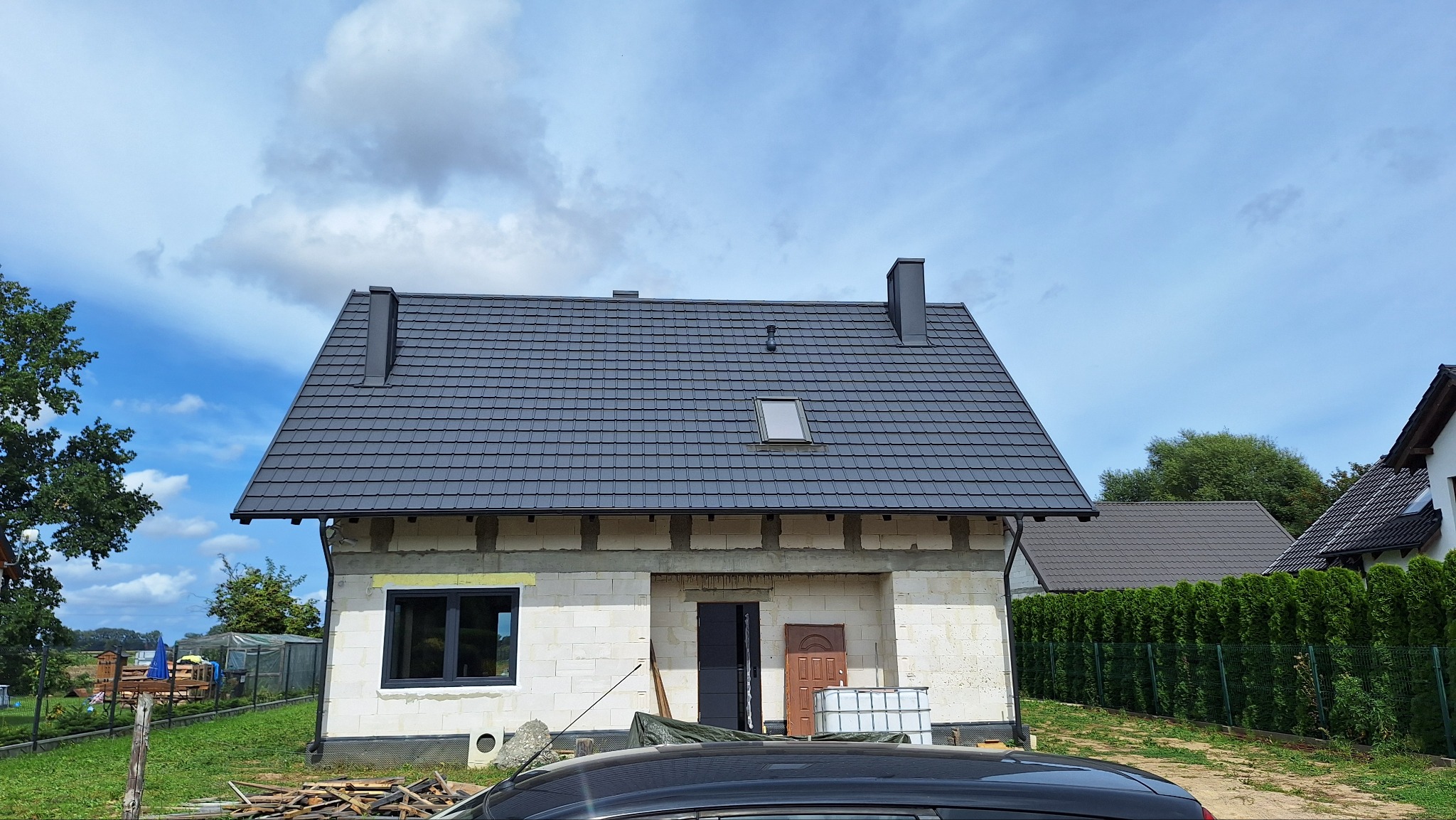 Nowy dom w trakcie budowy z ciemnoszarą dachówką, widoczne okno dachowe i komin, ściany z białego bloczka, częściowo ocieplone, na pierwszym planie fragment samochodu i stos drewna.