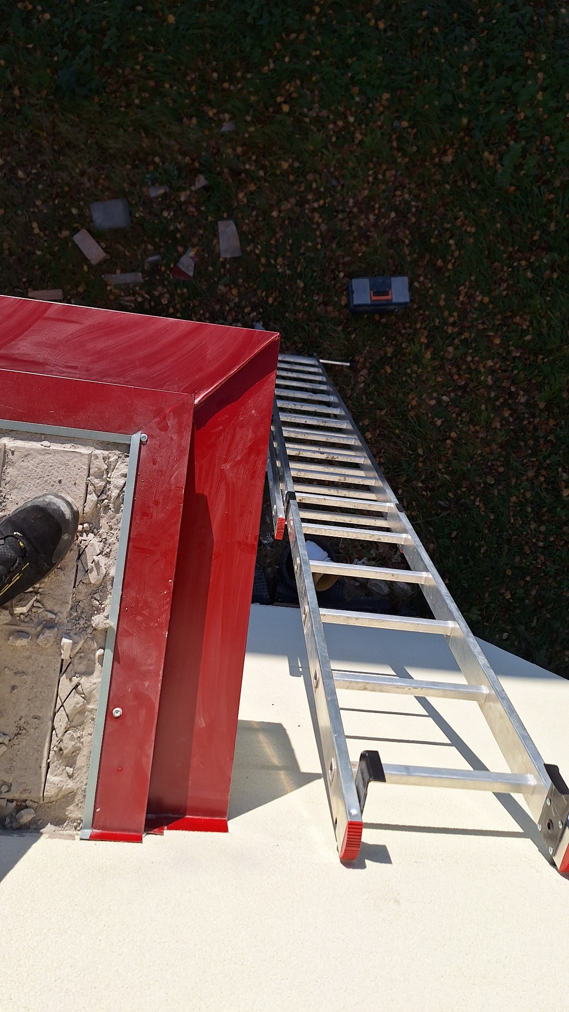 Widok z góry na fragment dachu z czerwoną obróbką blacharską, drabinę opartą o dach i narzędzia w tle na trawie.