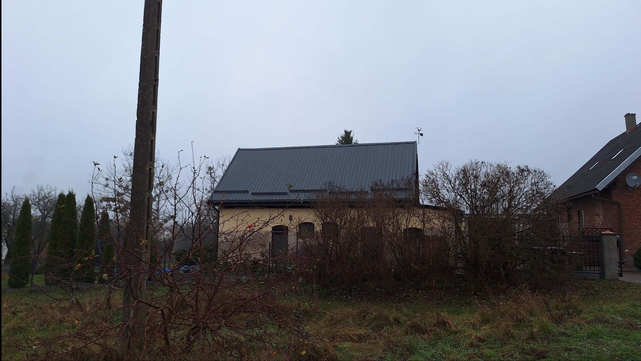 Dom jednorodzinny z nowym, ciemnoszarym dachem z blachy trapezowej, widok z poziomu gruntu, pochmurna pogoda, w tle widoczny słup elektryczny i sąsiedni budynek.