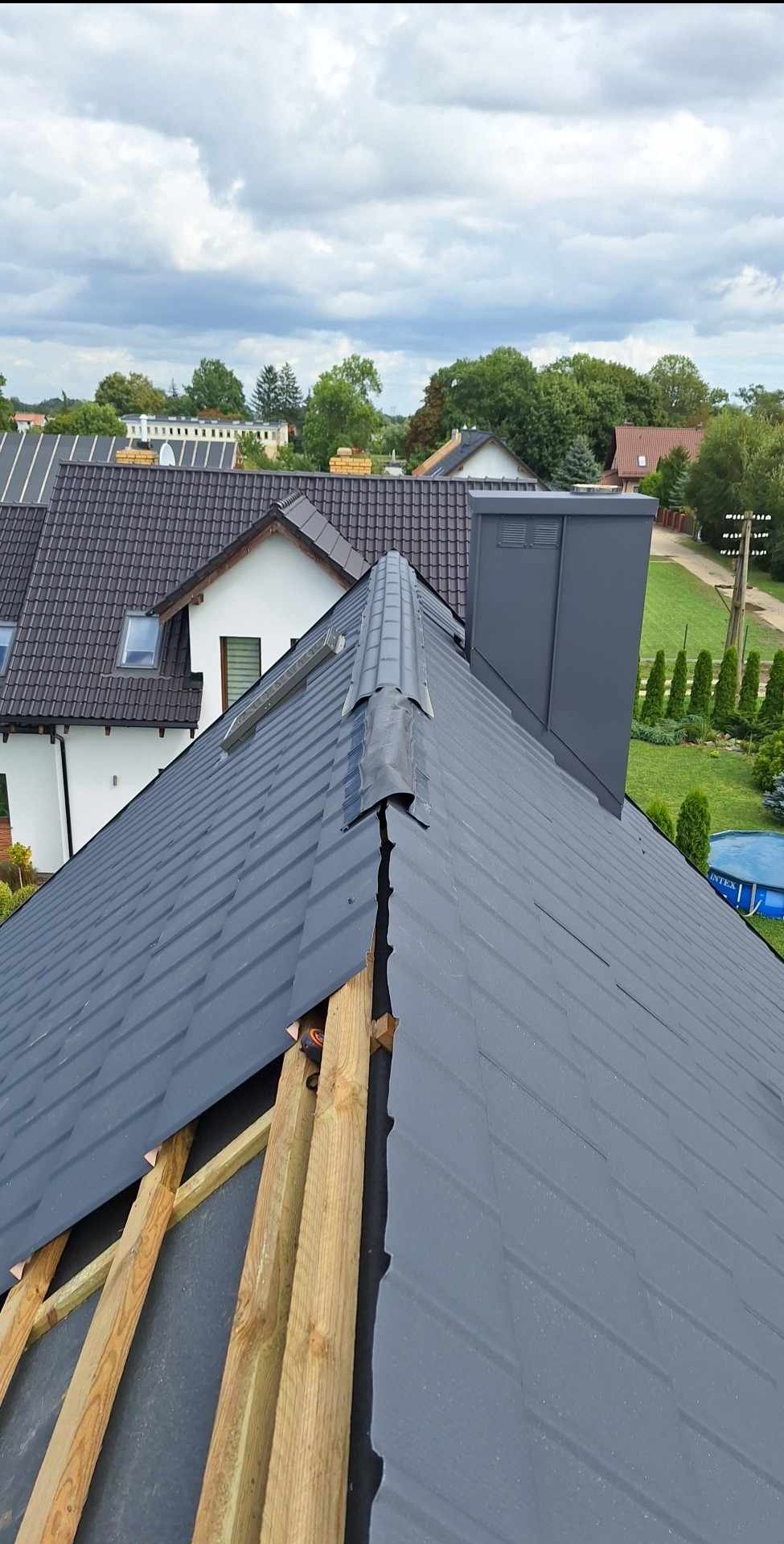 Szary dach z blachy trapezowej w trakcie montażu, widoczne drewniane łaty, w tle zabudowa jednorodzinna i zielone drzewa.