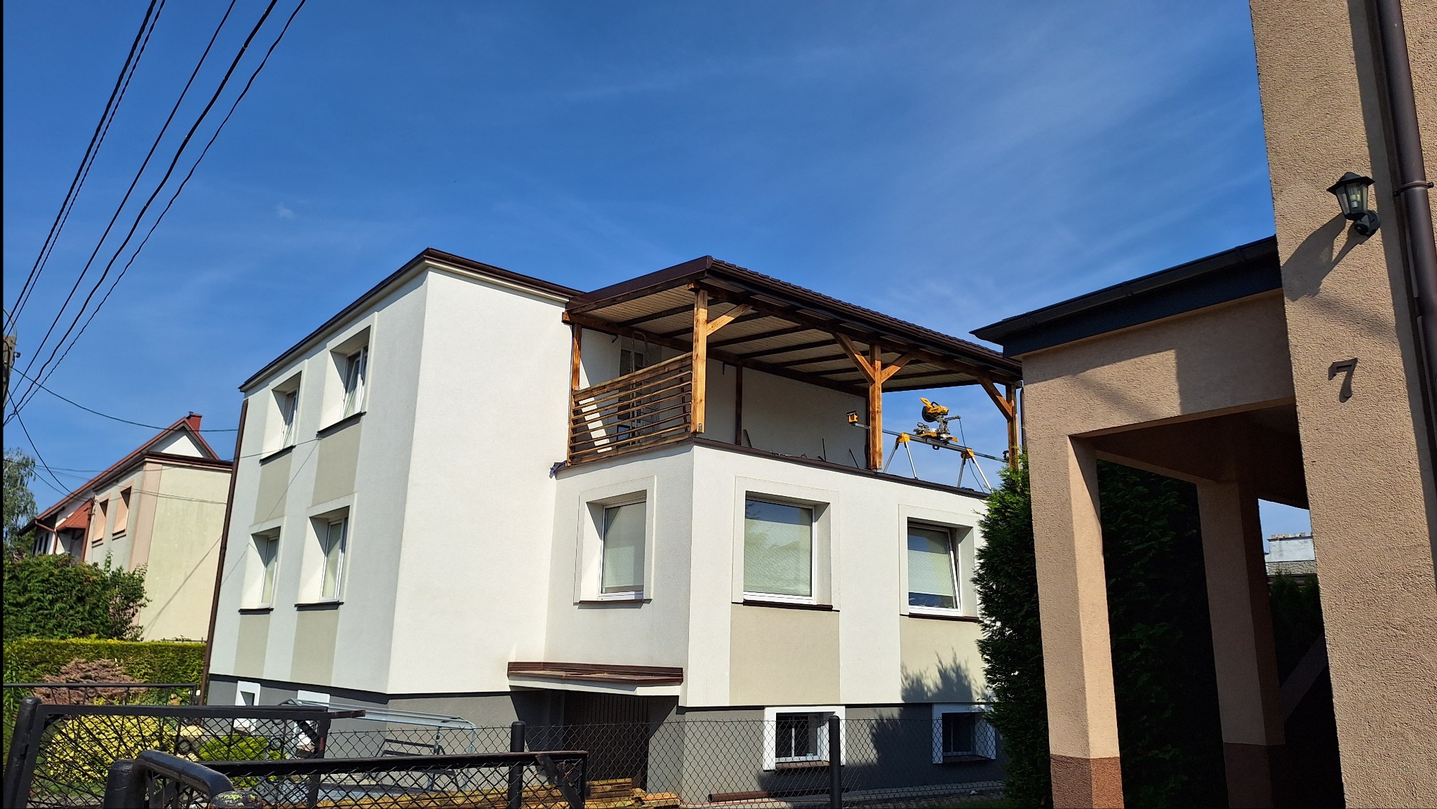Dwukondygnacyjny dom z drewnianą konstrukcją zadaszenia balkonu, widoczna piła ukosowa na stojaku, błękitne niebo w tle.