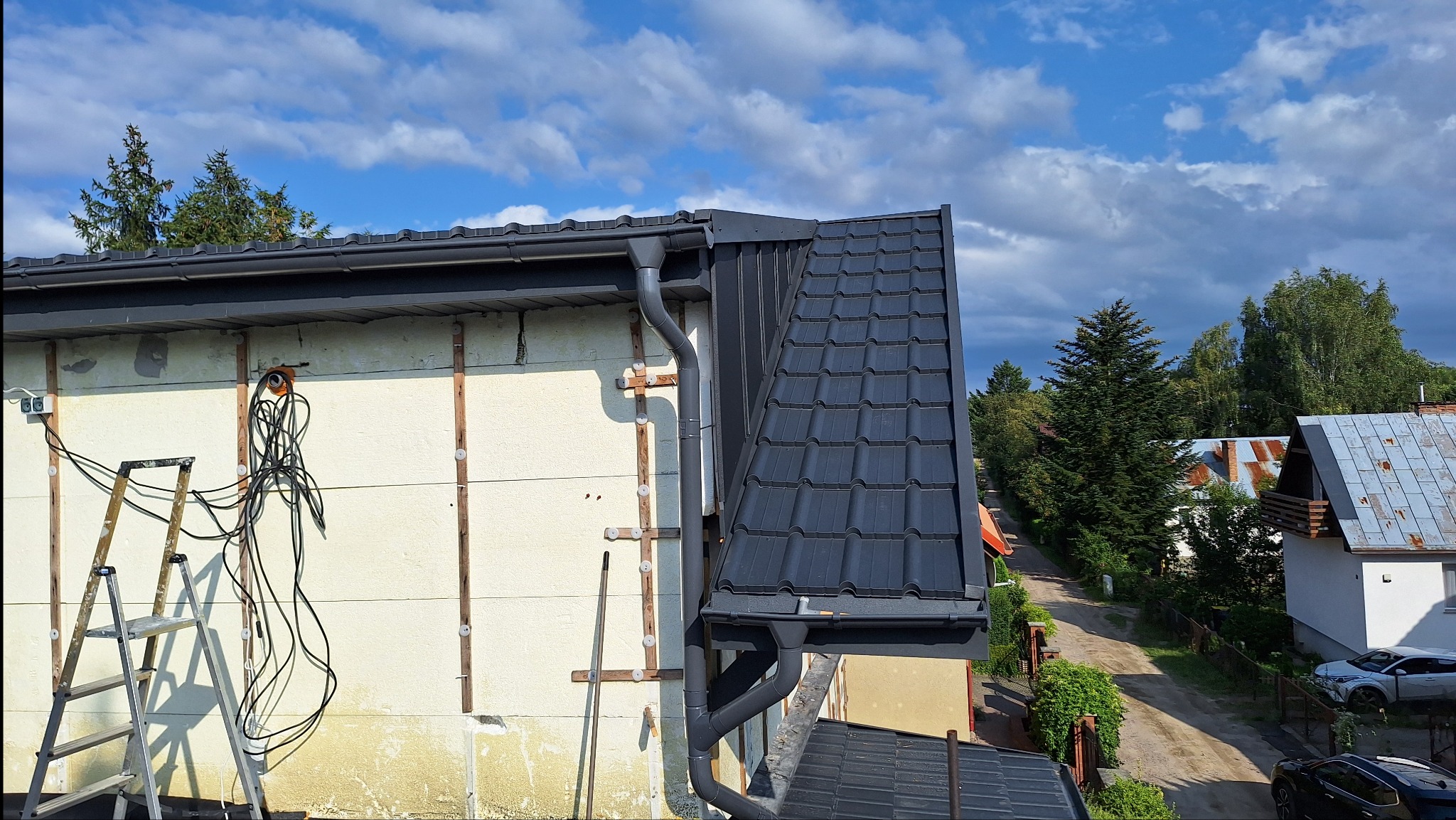 Fragment domu z nowym, grafitowym dachem z dachówki ceramicznej, widoczna rynna, ocieplenie ściany styropianem i ruszt drewniany, drabina oparta o ścianę, na tle błękitne niebo z chmurami...