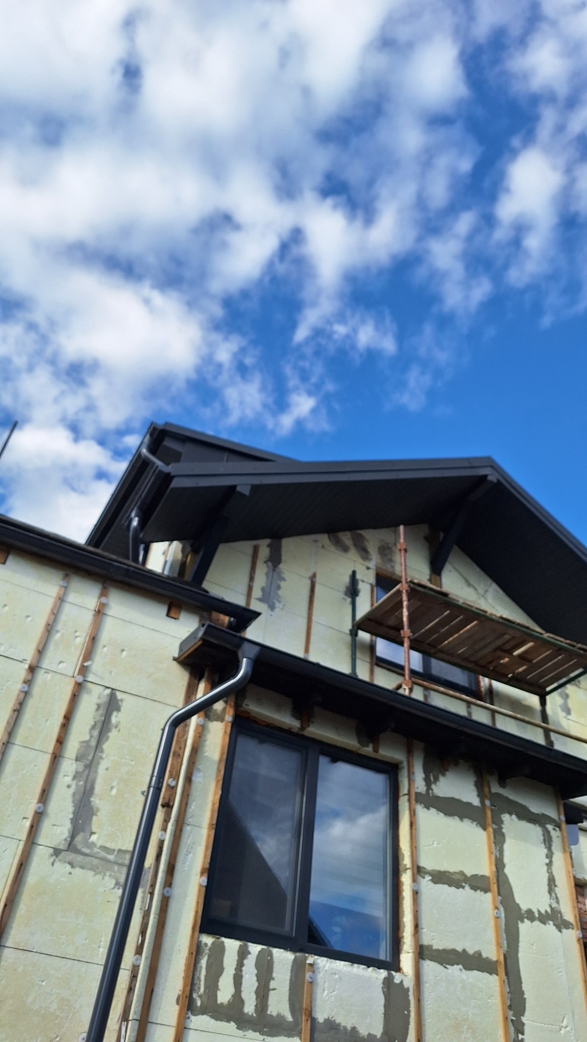 Dom w trakcie ocieplania, widoczne płyty izolacyjne na ścianach zewnętrznych, czarne rynny i fragment dachu, rusztowanie. Błękitne niebo z białymi chmurami w tle.