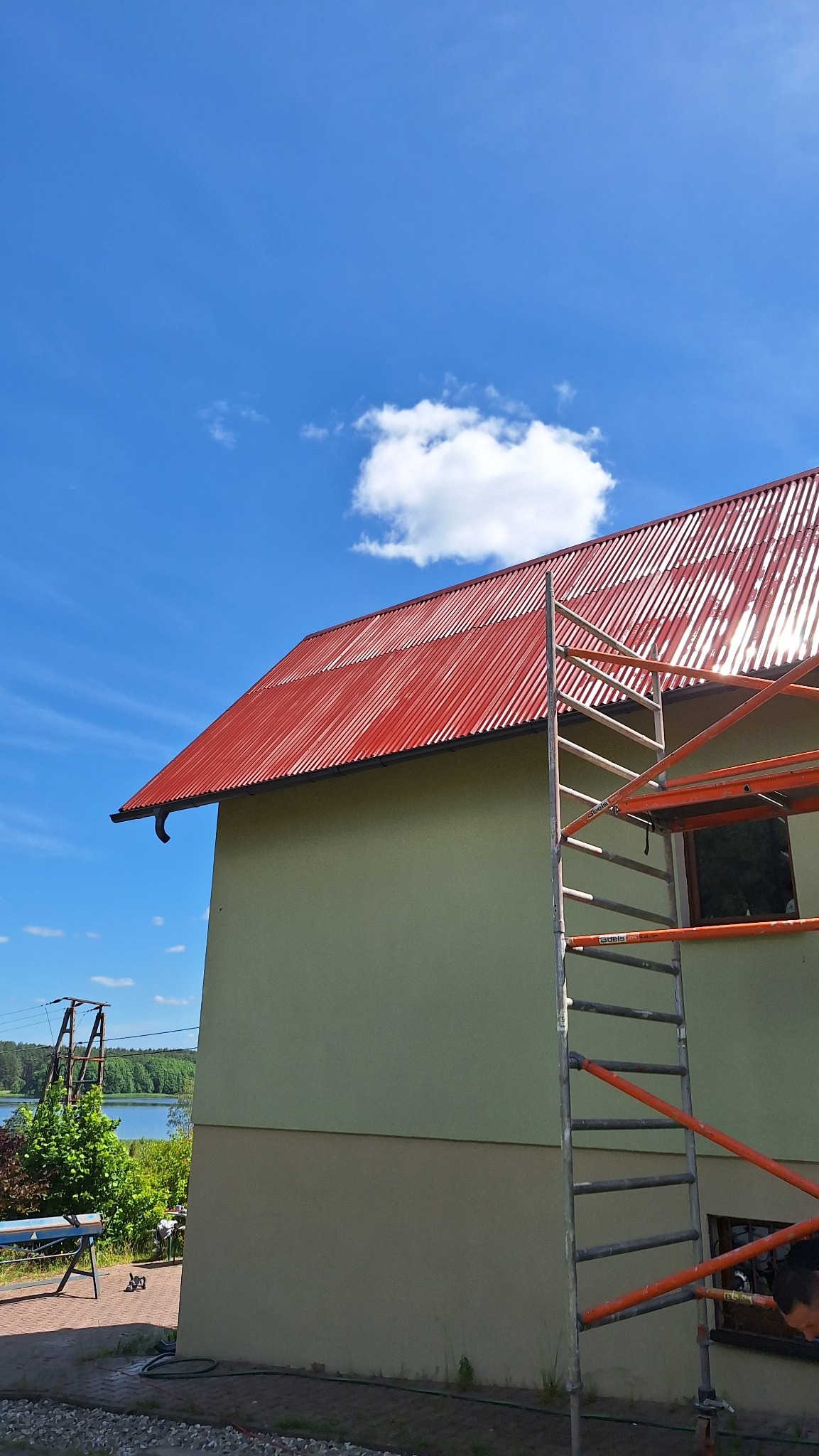 Czerwony, blaszany dach domu jednorodzinnego widziany z dołu, z częściowo widocznym rusztowaniem i fragmentem jeziora w tle.