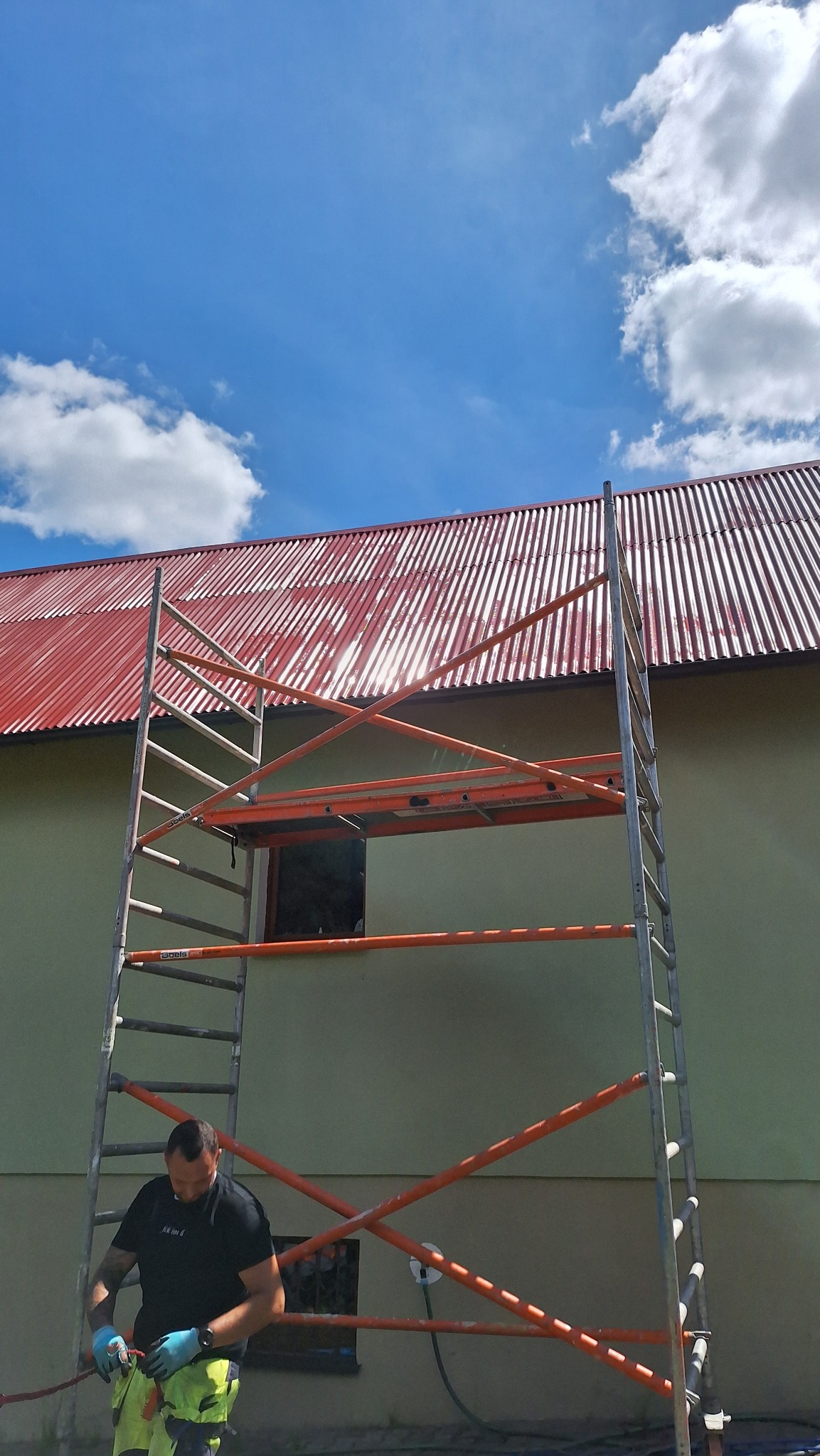 Pracownik w rękawicach przygotowuje się do pracy na rusztowaniu przed budynkiem z czerwonym dachem z blachy trapezowej, widoczne niebieskie niebo z chmurami.