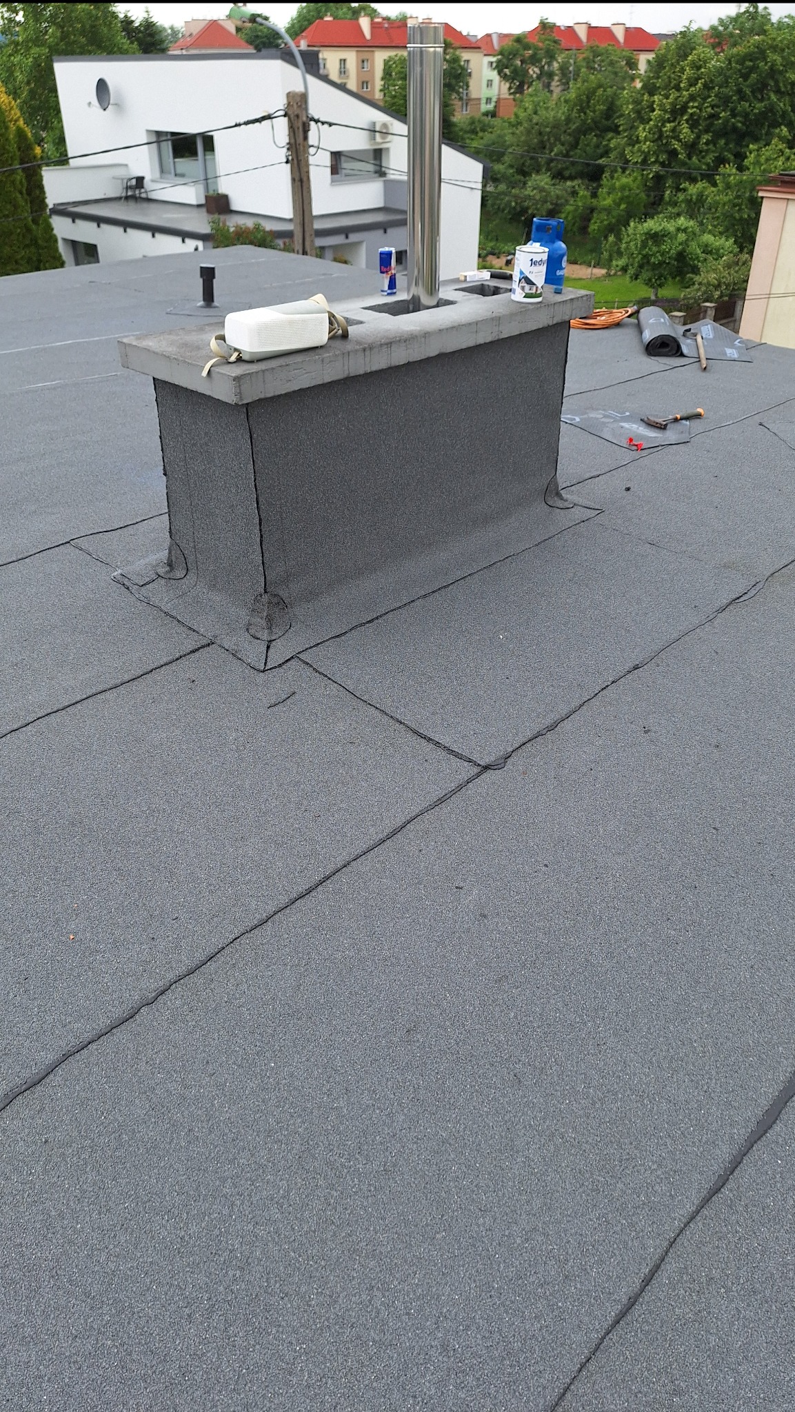 Szary, płaski dach pokryty papą, z kominem obłożonym papą i stalowym wkładem kominowym, widoczne narzędzia i materiały budowlane, tło stanowią budynki mieszkalne i zieleń.