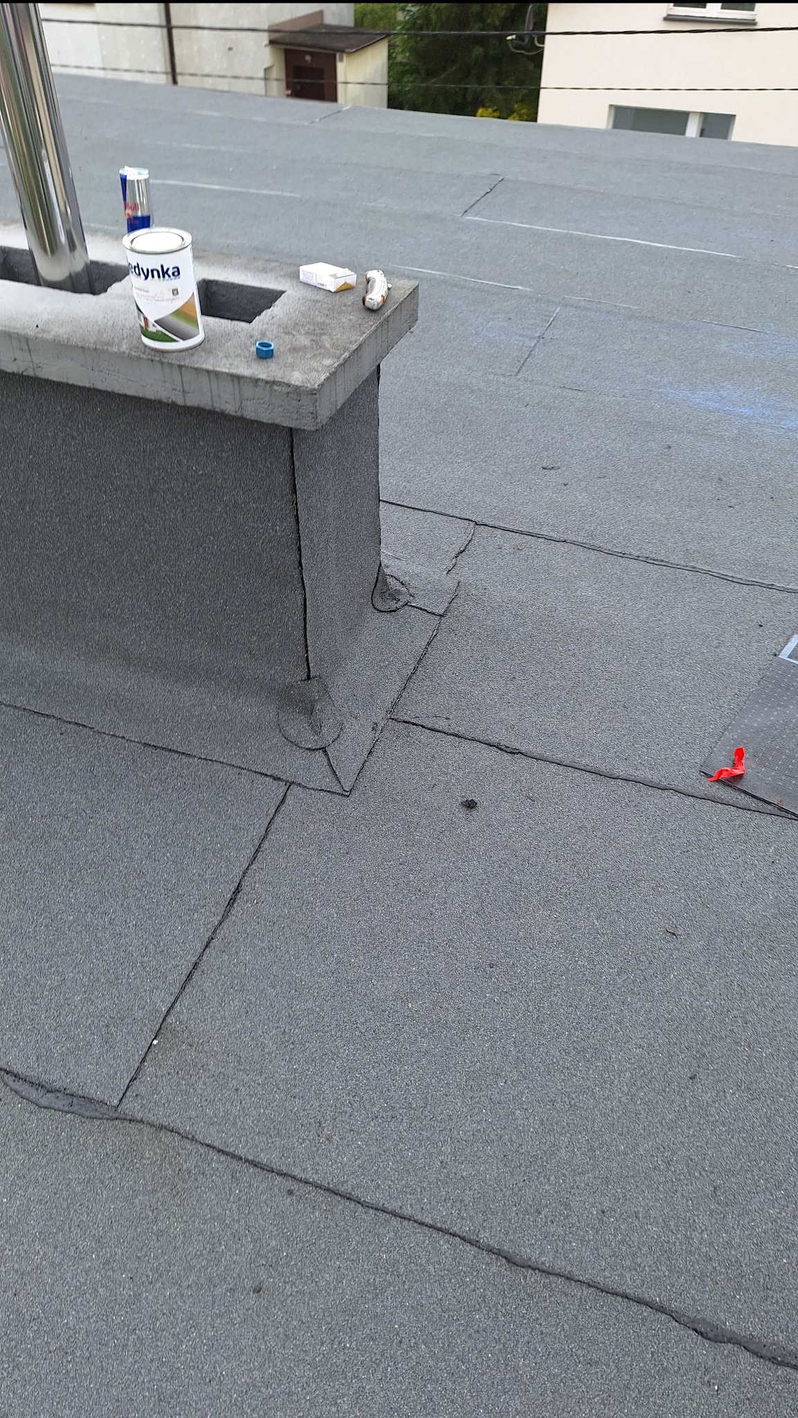 Płaski dach pokryty papą z widocznym kominem obłożonym papą, na którym leżą puszka farby, napój energetyczny, paczka papierosów i zapalniczka. Widoczne łączenia papy na dachu.