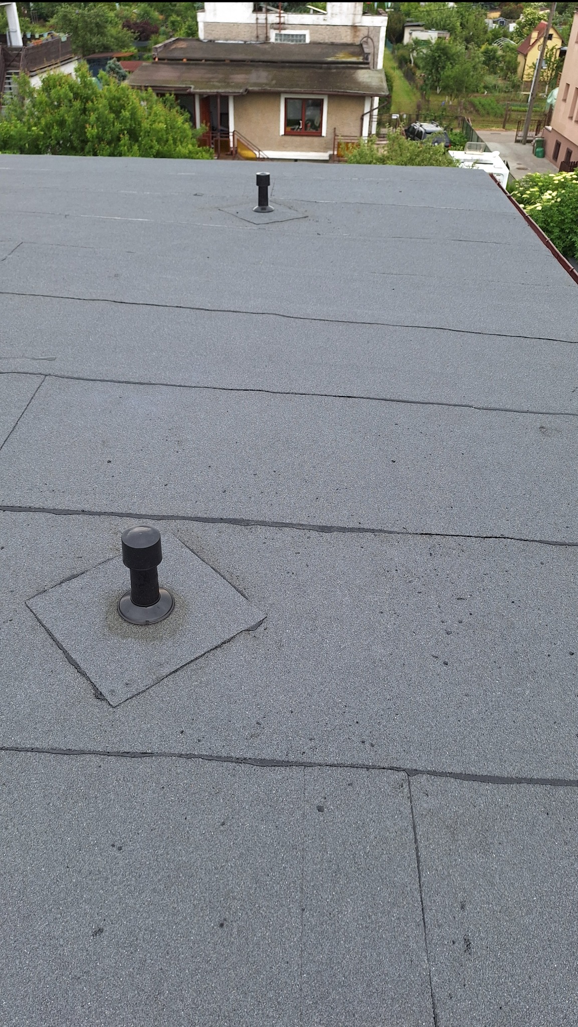 Szary, płaski dach pokryty papą termozgrzewalną w Bystrej z dwoma kominkami wentylacyjnymi. Widok z góry na budynki i zieleń w tle.