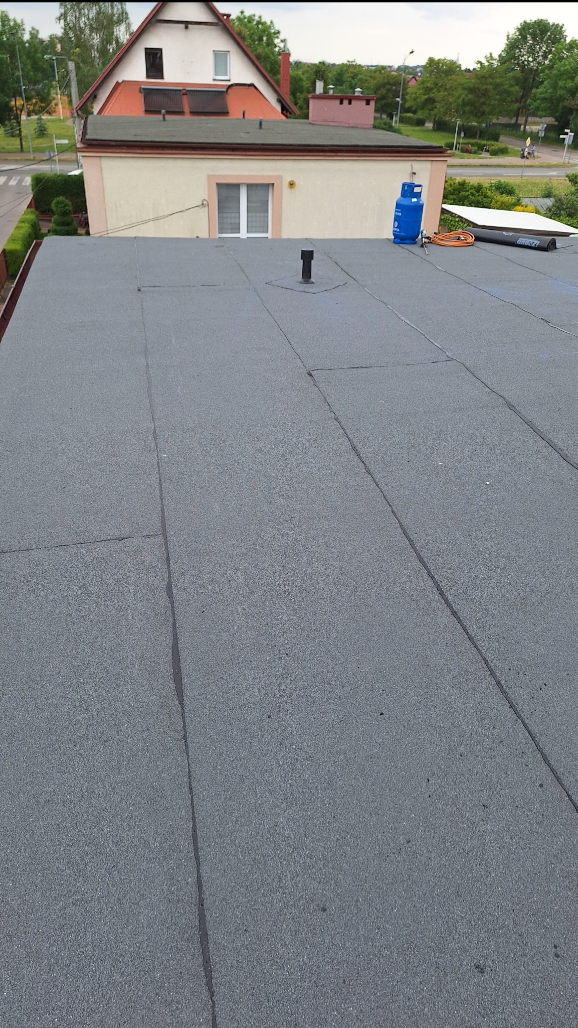 Szary, płaski dach pokryty papą termozgrzewalną z widoczną butlą gazową i zwiniętą rolką papy w tle.