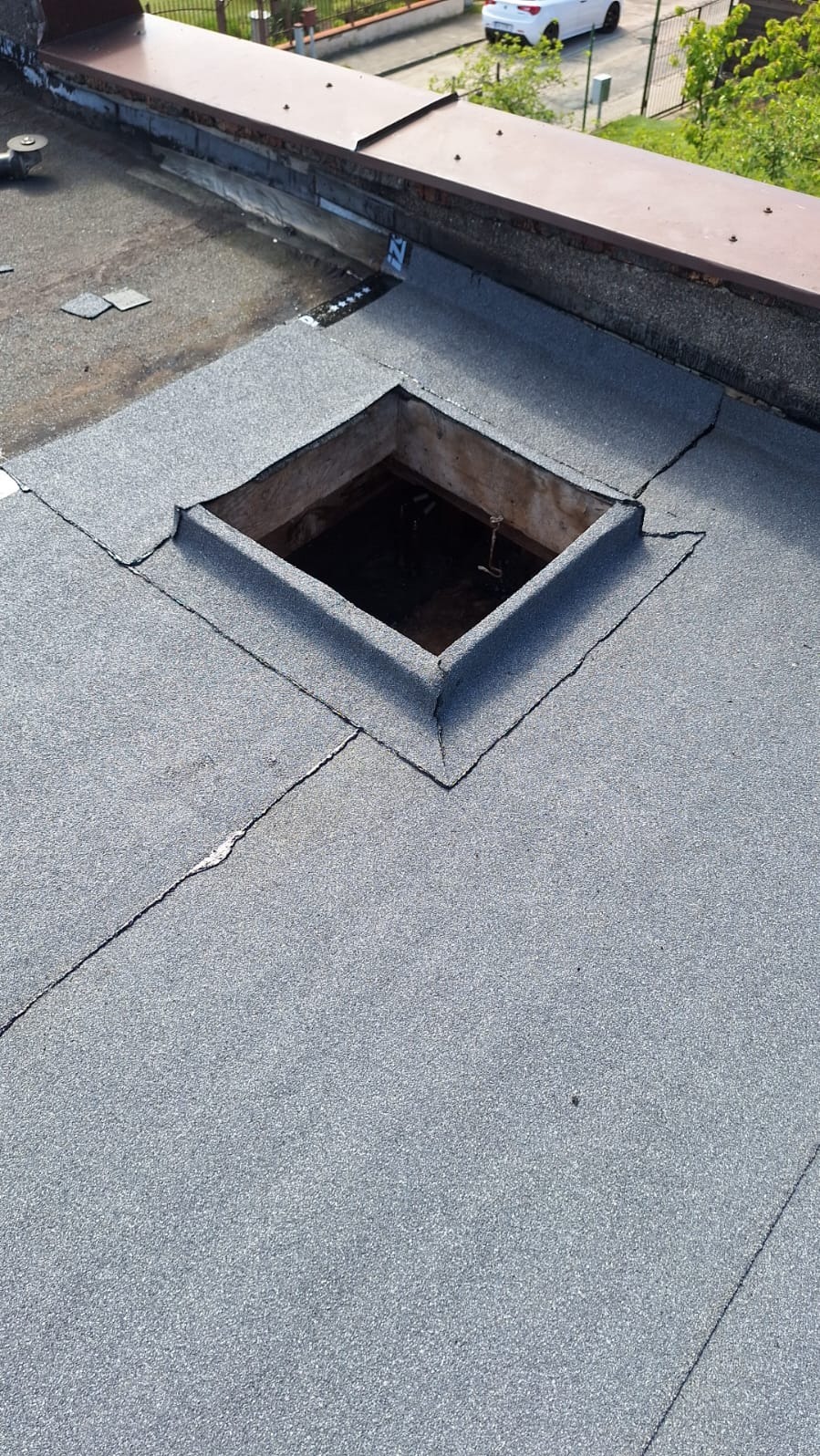 Płaski dach pokryty papą z kwadratowym otworem wentylacyjnym, widoczne łączenia papy i fragment obróbki blacharskiej przy krawędzi dachu.