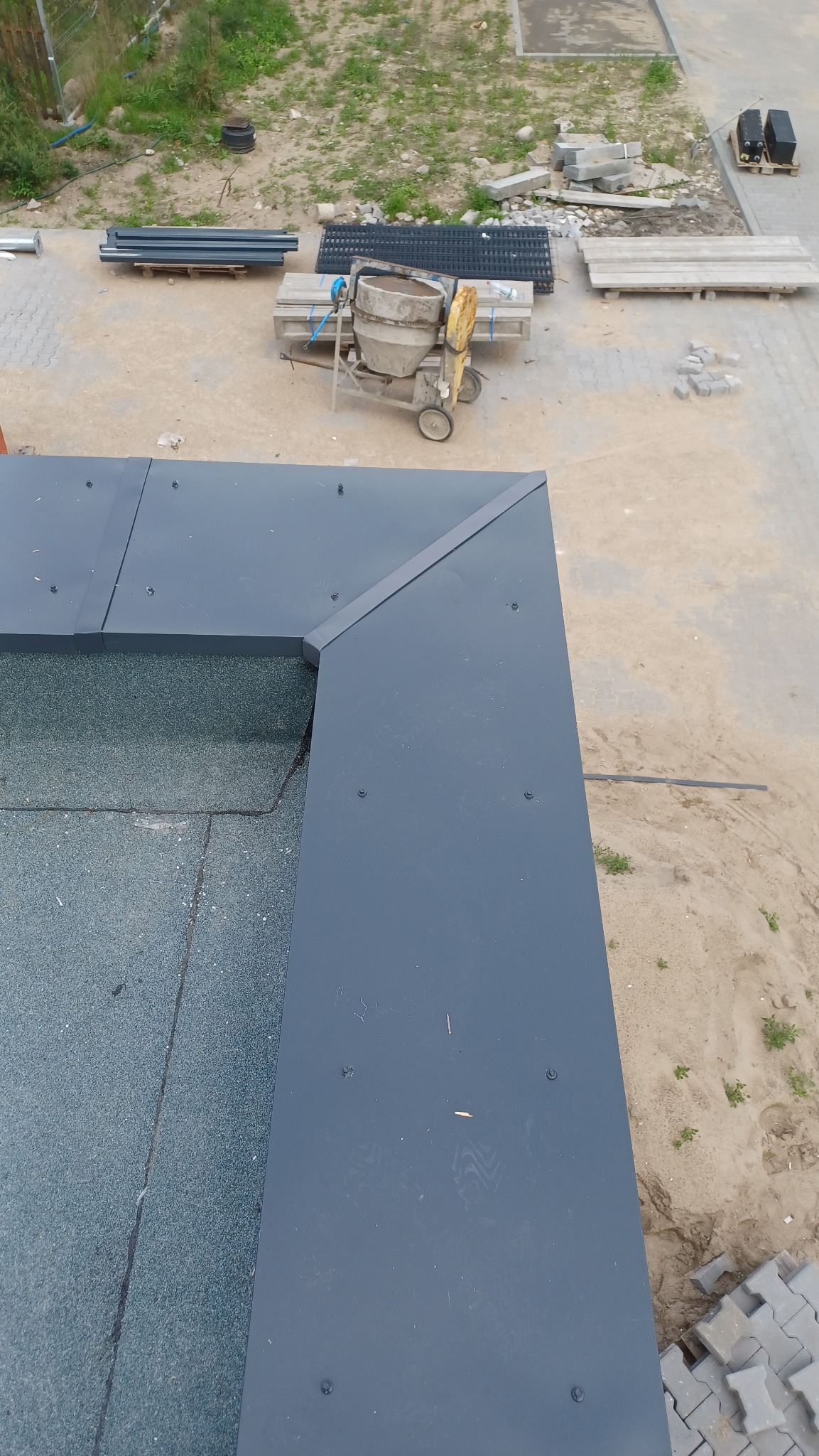 Widok z góry na fragment dachu z nową, grafitową obróbką blacharską na krawędziach, z widocznymi śrubami mocującymi; w tle teren budowy z betoniarką i materiałami.