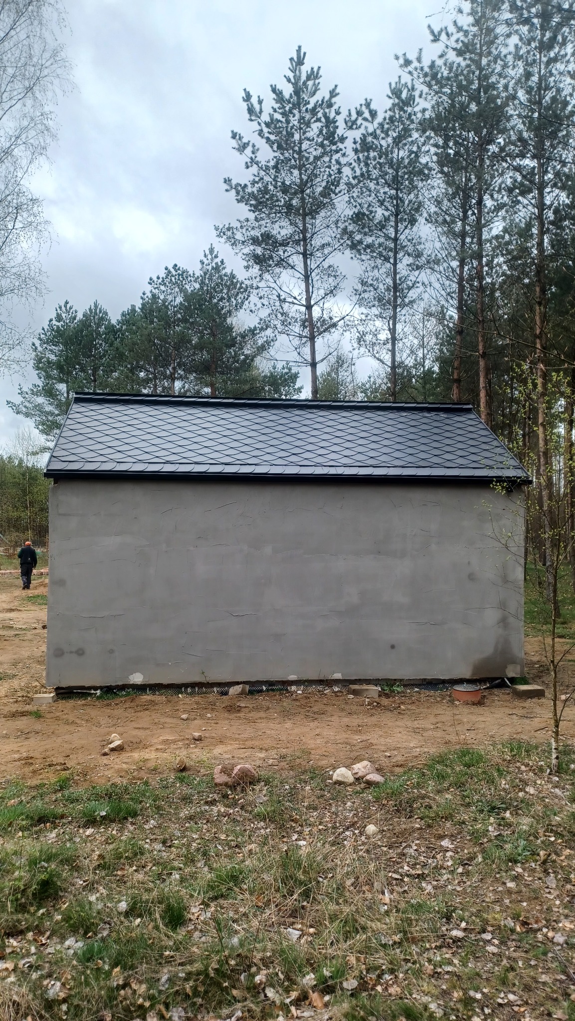 Niewielki, szary budynek z nowym, czarnym dachem z dachówki bitumicznej w kształcie rybiej łuski, widoczny na tle lasu iglastego i pochmurnego nieba. W tle sylwetka człowieka w ciemnym ubraniu.