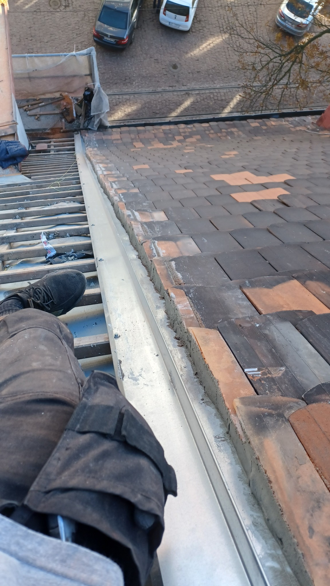 Widok z góry na fragment dachu z częściowo ułożoną dachówką, widoczna metalowa obróbka blacharska i noga robotnika w czarnym obuwiu roboczym, w tle zabudowa miejska.