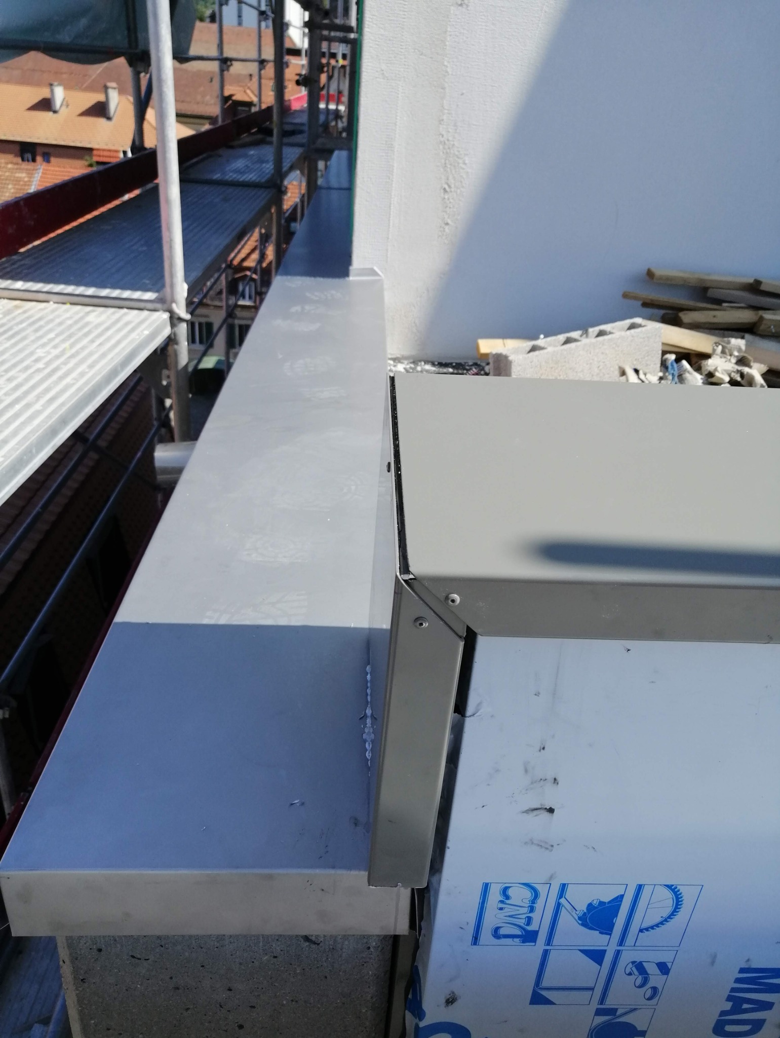 Szare, metalowe obróbki blacharskie dachu, widoczne z rusztowania, z fragmentem białej ściany w tle i elementami konstrukcyjnymi dachu w oddali.