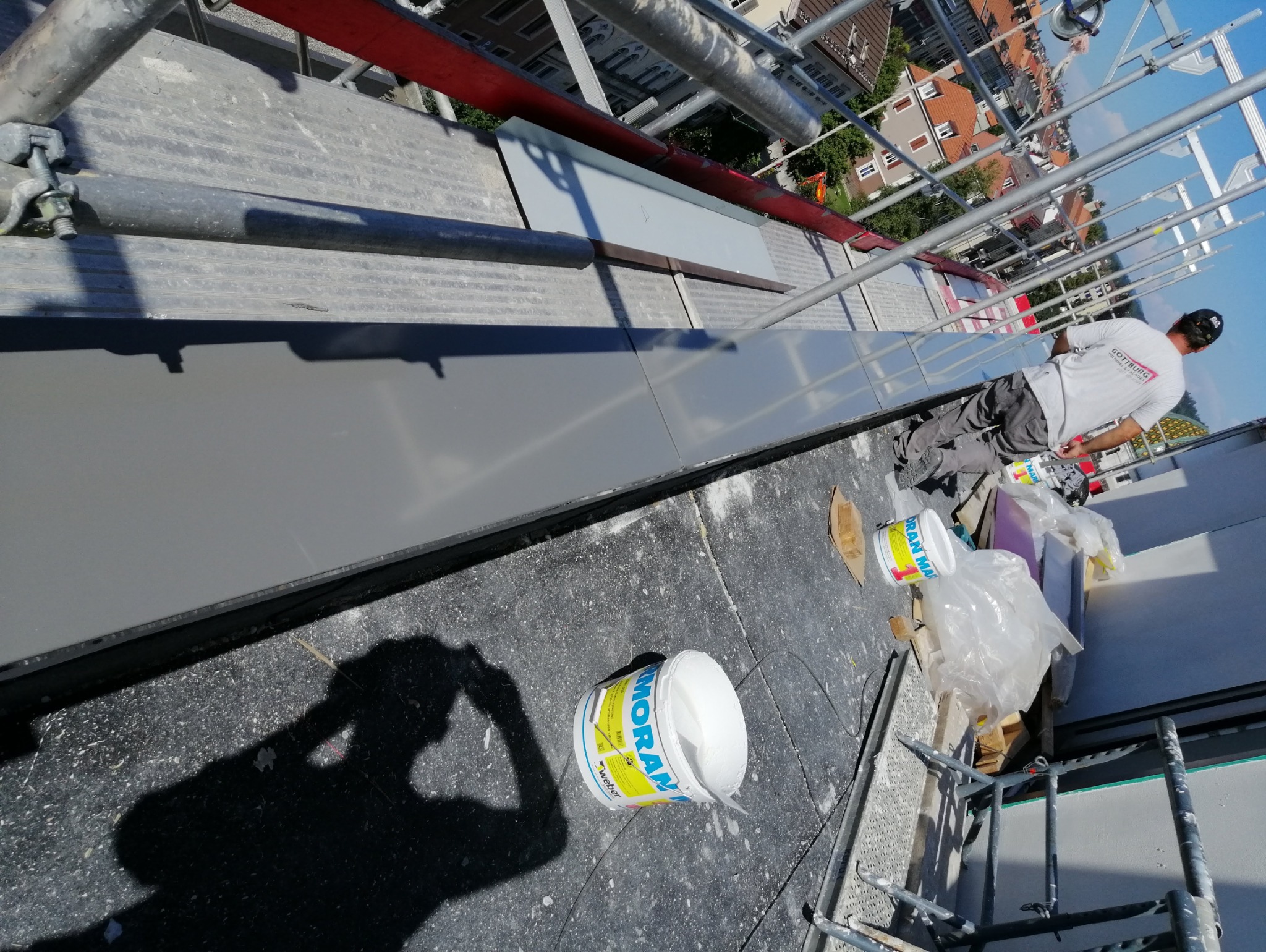 Praca na wysokości: blacharz na rusztowaniu przy montażu szarej obróbki blacharskiej na dachu, widoczne puszki z farbą i narzędzia, cień fotografa w lewym dolnym rogu.