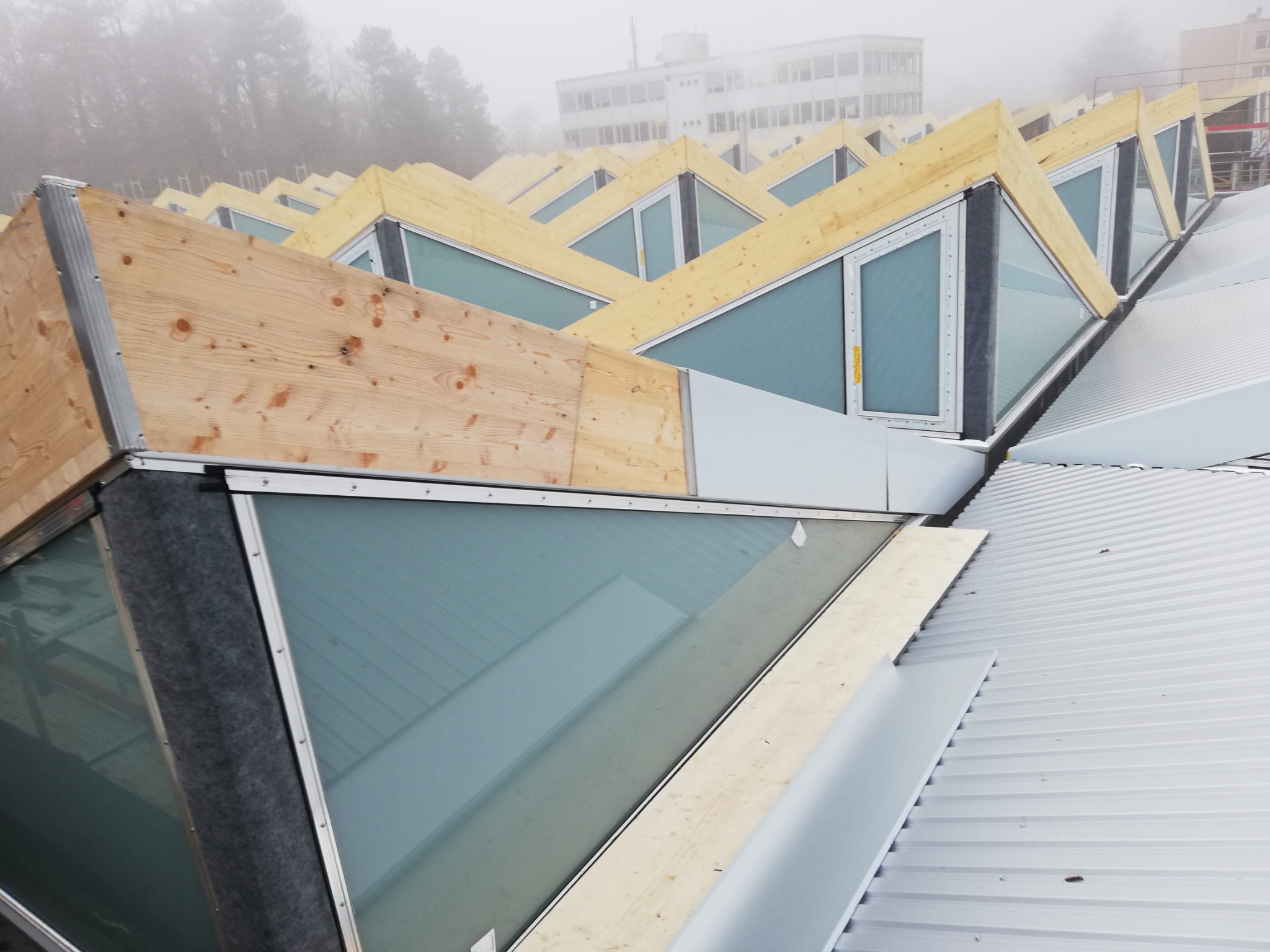 Montaż okien dachowych w konstrukcji dachu z blachy trapezowej, widoczne drewniane elementy konstrukcyjne i uszczelnienie, budynek w tle we mgle.