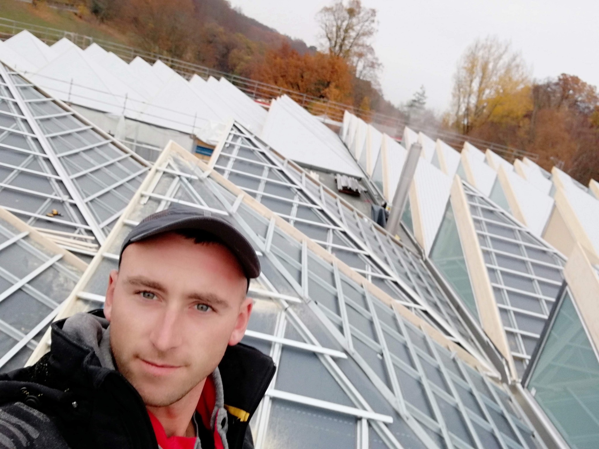 Selfie dekarza na tle szkieletu dachu z licznymi przeszkleniami, widoczne jesienne drzewa w tle.