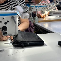 HATEX SPÓŁKA Z OGRANICZONĄ ODPOWIEDZIALNOŚCIĄ - Szwalnia: pracownik szyje czarną kosmetyczkę na maszynie przemysłowej. W tle inne stanowiska pracy i osoby przy maszynach. Oświetlenie lampą punktową.