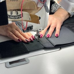 HATEX SPÓŁKA Z OGRANICZONĄ ODPOWIEDZIALNOŚCIĄ - Praca przy maszynie do szycia: zszywanie czarnego, grubego materiału z rzepami. Widoczne dłonie, igła maszyny i fragmenty materiału. Oświetlenie lampą.