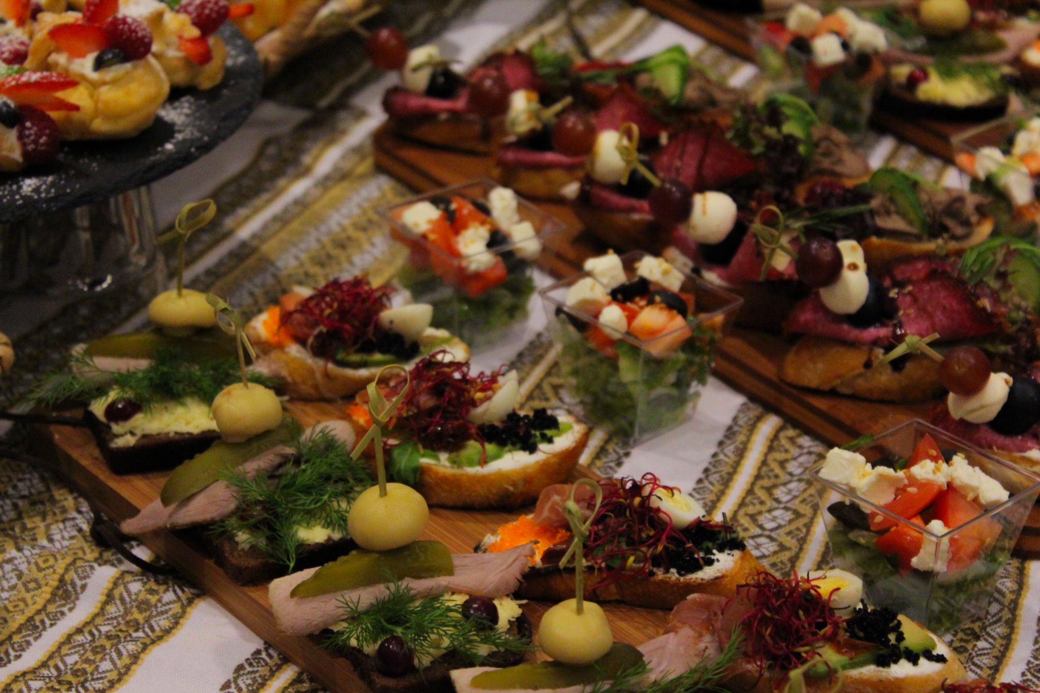 Różnorodne kanapeczki i sałatki w plastikowych pojemnikach, ułożone na drewnianych deskach z dekoracyjnymi pikami, prezentowane na wzorzystym obrusie.