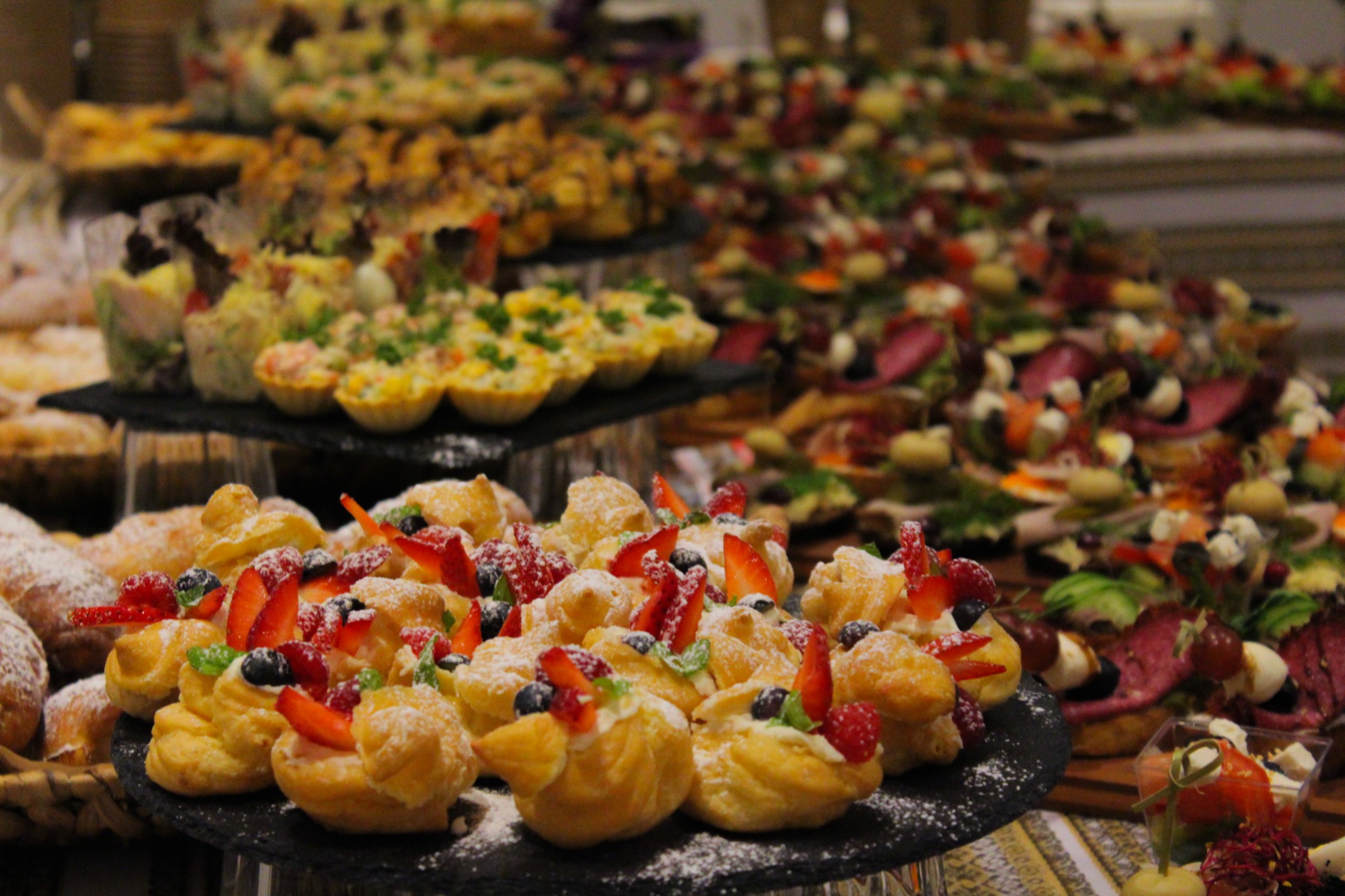 Bogato zastawiony stół z różnorodnymi przekąskami: mini-ptysie z kremem i owocami, tartaletki z pastami, kanapeczki z wędlinami i warzywami, ułożone na kilku poziomach.