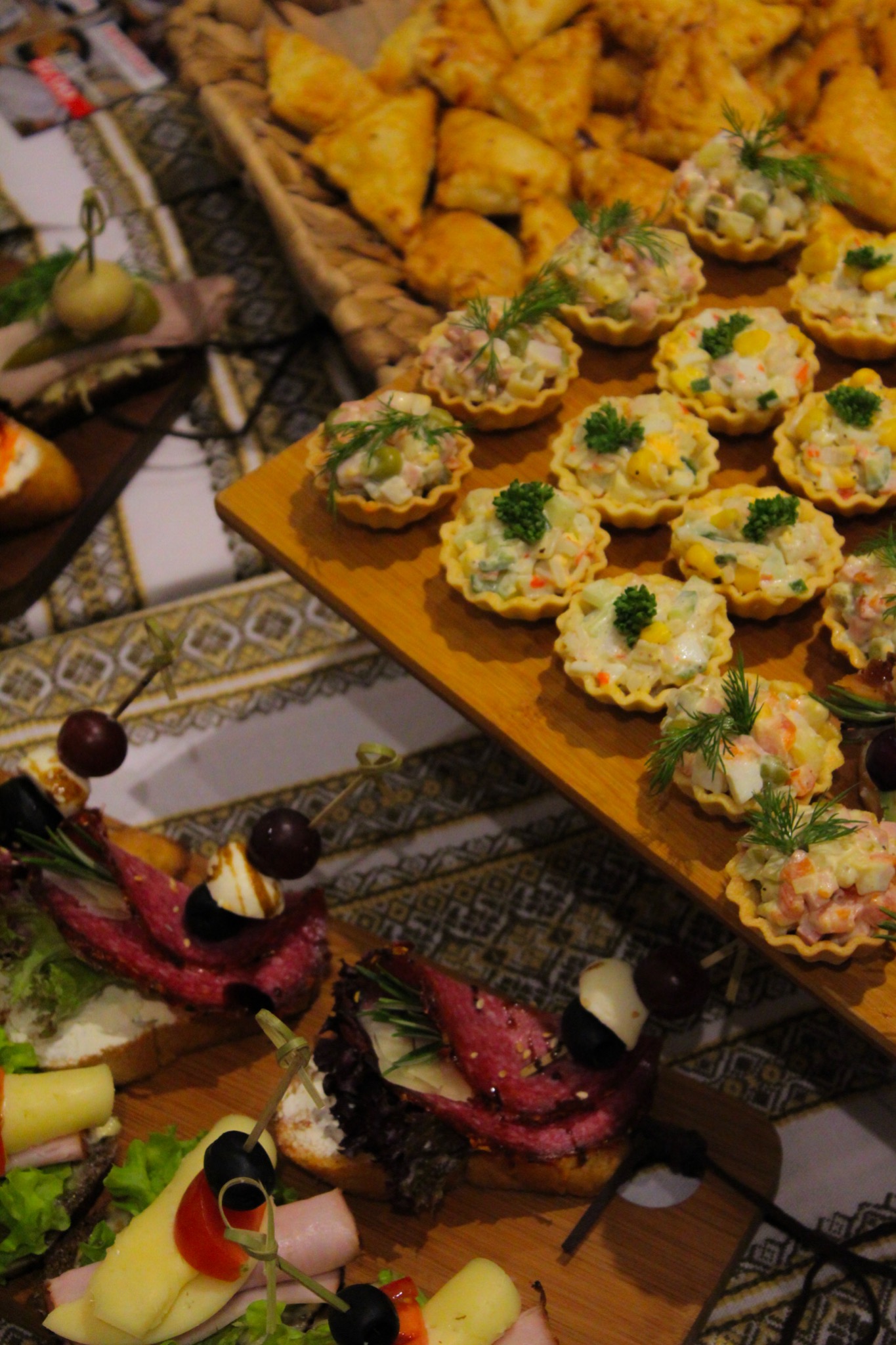 Różnorodny zestaw przekąsek na drewnianych deskach: tartaletki z sałatką, kanapeczki z wędliną i serem, oraz pierożki w wiklinowym koszyku, ułożone na wzorzystym obrusie.