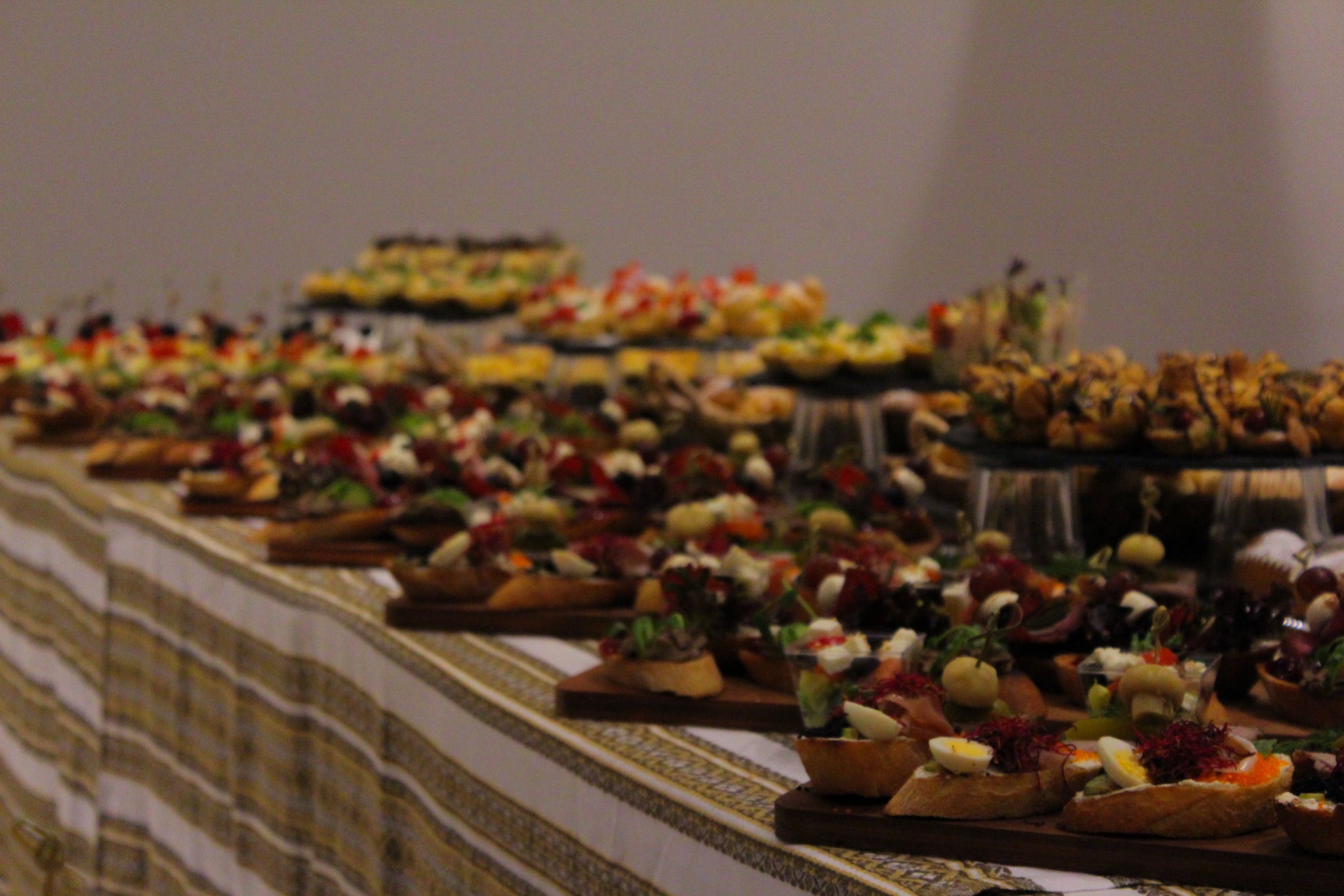 Stół cateringowy z różnorodnymi przekąskami na drewnianych deskach i podwyższeniach, w tle szara ściana, oświetlenie punktowe.