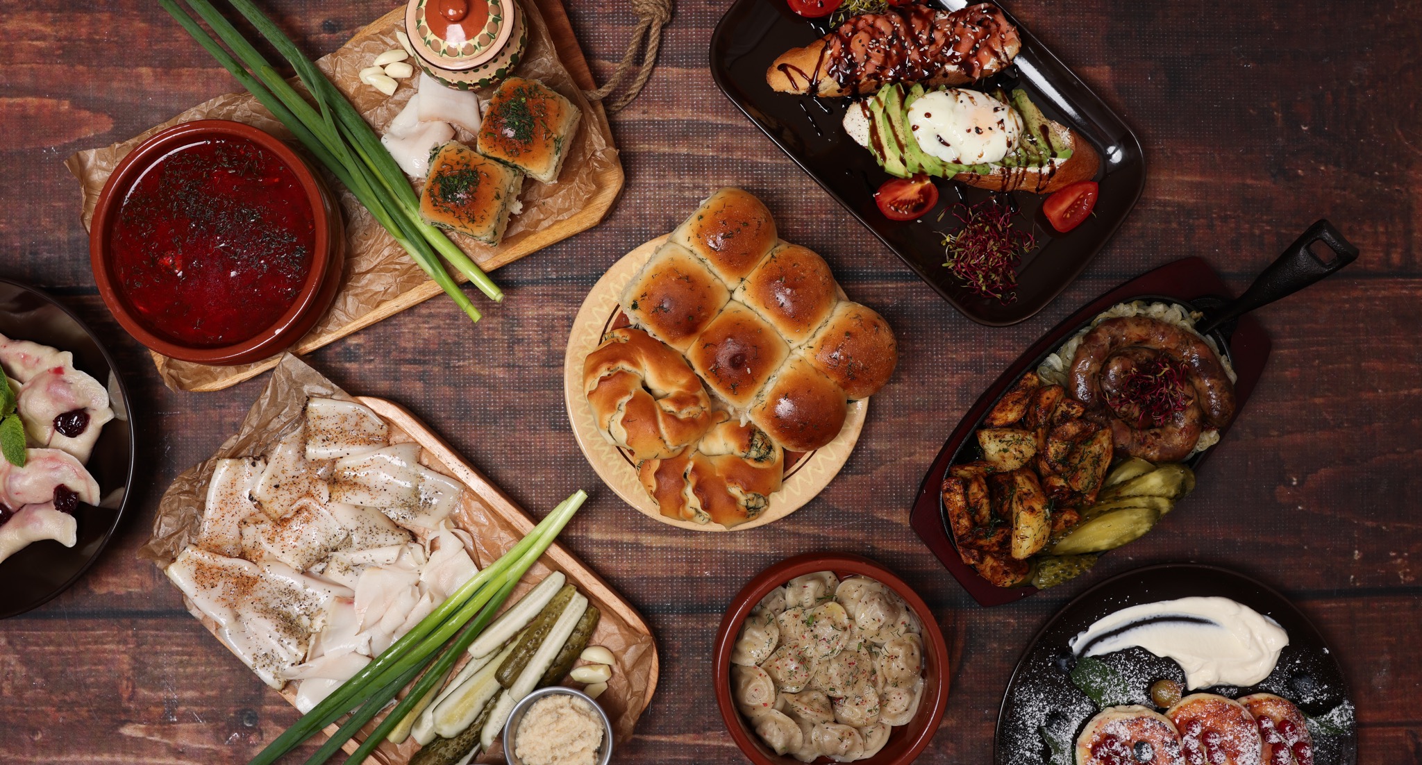 Bogaty stół z różnorodnymi daniami kuchni wschodnioeuropejskiej: barszcz czerwony w glinianej misce, pierogi z wiśniami, słonina wędzona, pieczone pierożki z farszem, bułeczki drożdżowe, pierogi...