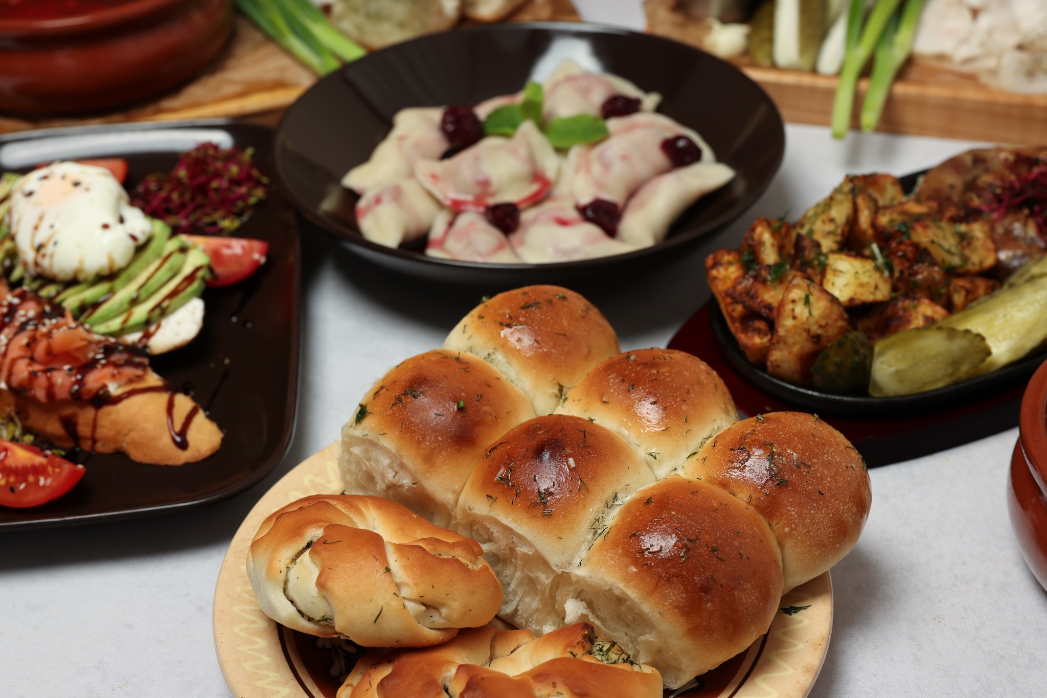 Urozmaicony stół z różnymi daniami: pierogi z wiśniami, mini bułeczki posypane koperkiem, grzanki z awokado i jajkiem w koszulce oraz pieczone ziemniaki z ziołami, całość sfotografowana z bliska.