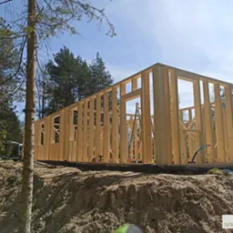 Wznoszenie drewnianej konstrukcji domu w systemie szkieletowym na fundamencie, widoczne pionowe i poziome elementy ramy, drabina oparta o ścianę, w tle drzewa i błękitne niebo.