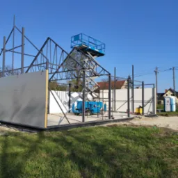 Montaż hali: stalowa konstrukcja szkieletowa z częściowo zamontowanymi szarymi panelami ściennymi, podnośnik koszowy wewnątrz, widoczna przenośna toaleta Toi Toi w tle, słoneczny dzień.