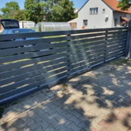 Nowoczesna przesuwna brama wjazdowa z poziomych profili, widok z zewnątrz na tle domu z ogrodem i samochodem z otwartym bagażnikiem.