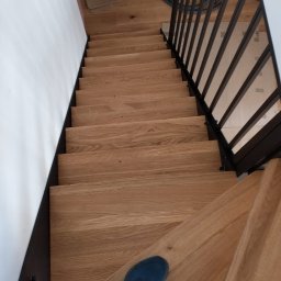 STENS SPÓŁKA Z OGRANICZONĄ ODPOWIEDZIALNOŚCIĄ - Widok z góry na drewniane schody z czarną metalową balustradą; w tle widoczna osoba z odkurzaczem przemysłowym; na pierwszym planie stopa w niebieskim bucie.