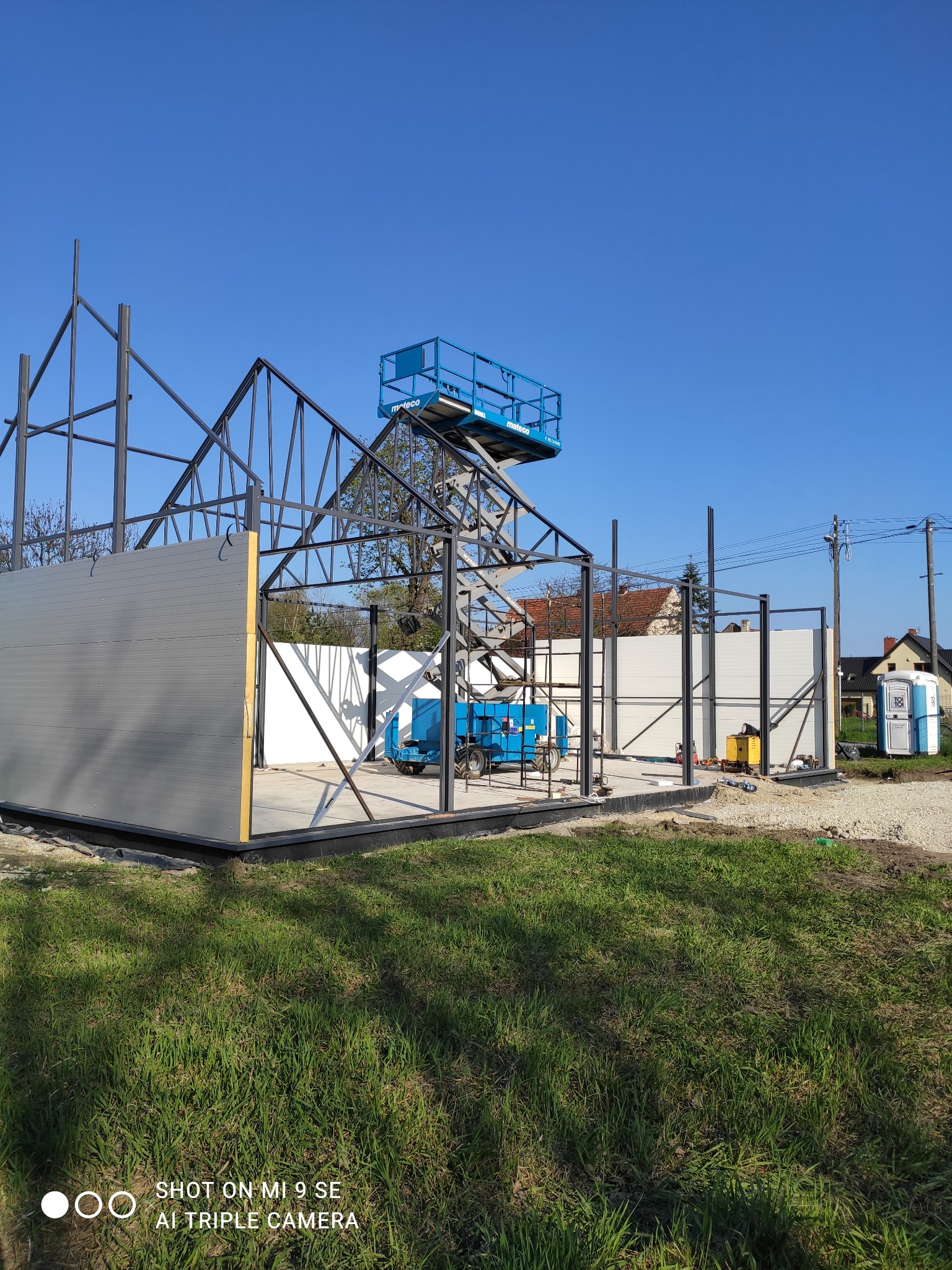 Montaż hali: stalowa konstrukcja szkieletowa z częściowo zamontowanymi szarymi panelami ściennymi, podnośnik koszowy wewnątrz, widoczna przenośna toaleta Toi Toi w tle, słoneczny dzień.