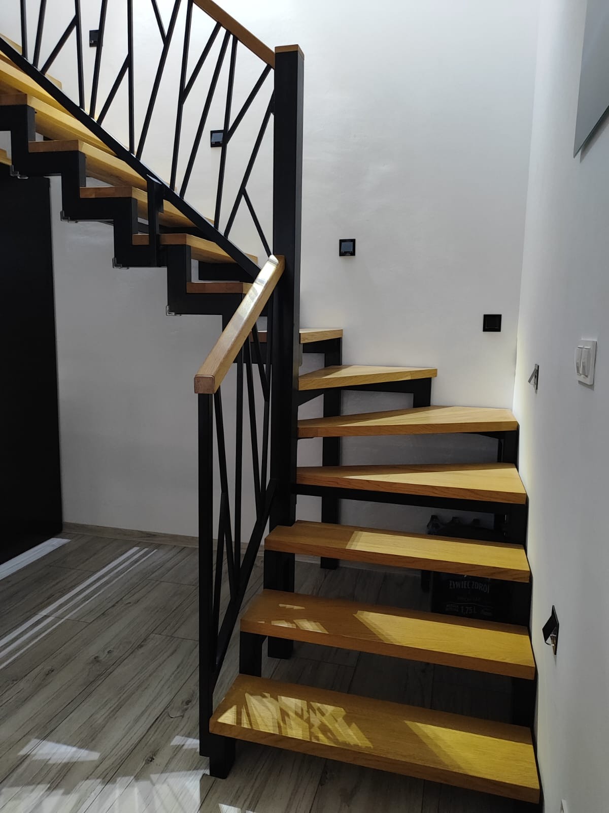 Nowoczesne schody metalowe z drewnianymi stopniami i balustradą z geometrycznym wzorem, oświetlone światłem dziennym, widoczne czarne włączniki na białej ścianie.