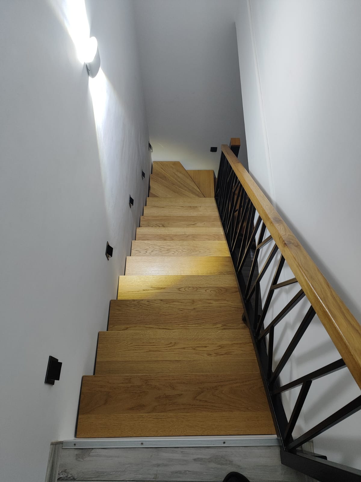 Wnętrze domu: widok z góry na drewniane schody z czarną, geometryczną balustradą i oświetleniem punktowym na białej ścianie.