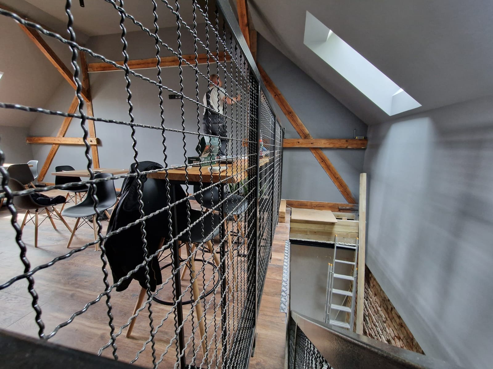 Klatka schodowa z metalową siatką, widok z góry na dół, ukazujący biuro na poddaszu z drewnianymi belkami i oknem dachowym.
