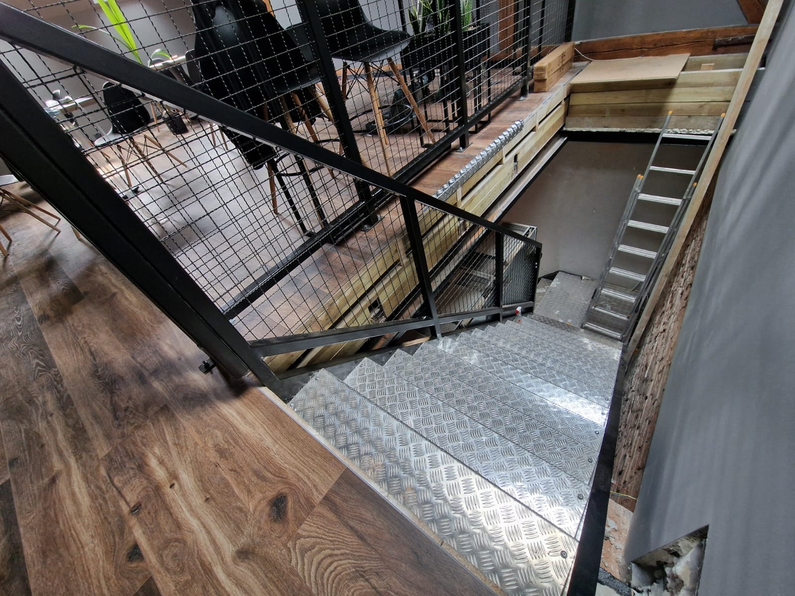 Wnętrze z metalowymi schodami o antypoślizgowej powierzchni, zabezpieczonymi czarną, siatkowaną balustradą. Widoczne drewniane wykończenie podestu oraz drabina oparta o ścianę w tle.