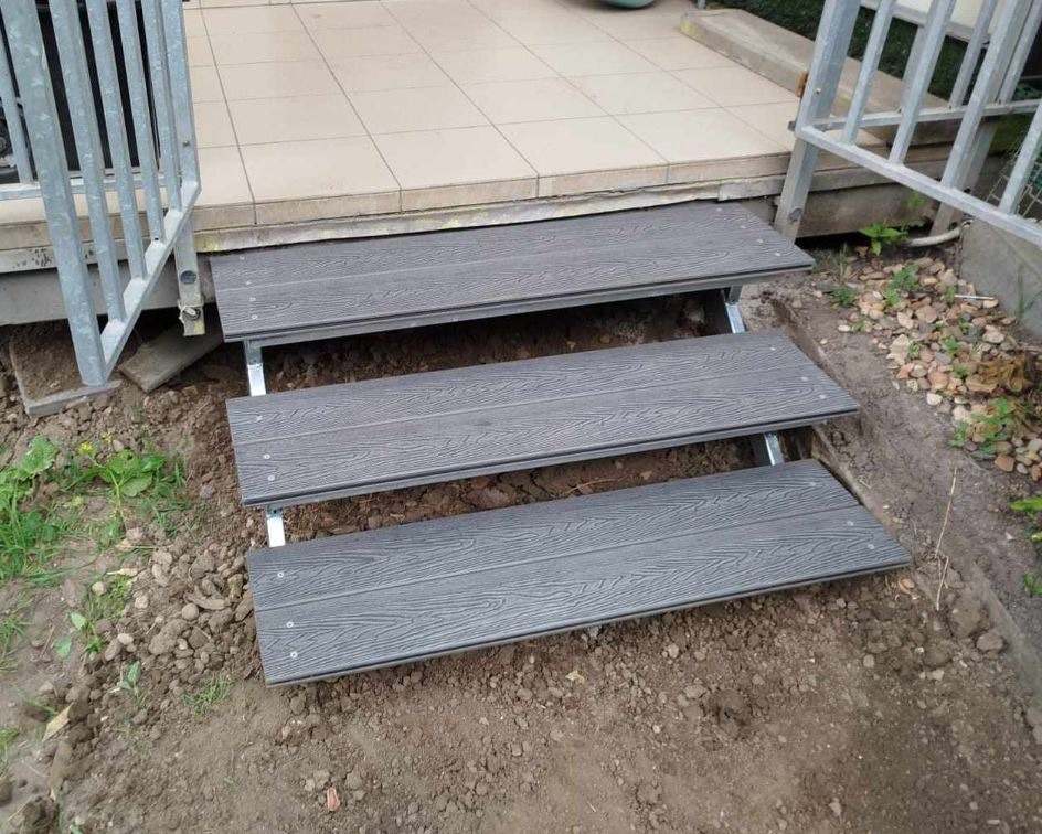 Trzy stopnie schodowe z kompozytu drewnopodobnego, zamontowane na metalowych wspornikach, prowadzące do wejścia z jasnymi płytkami, widoczne elementy balustrady.