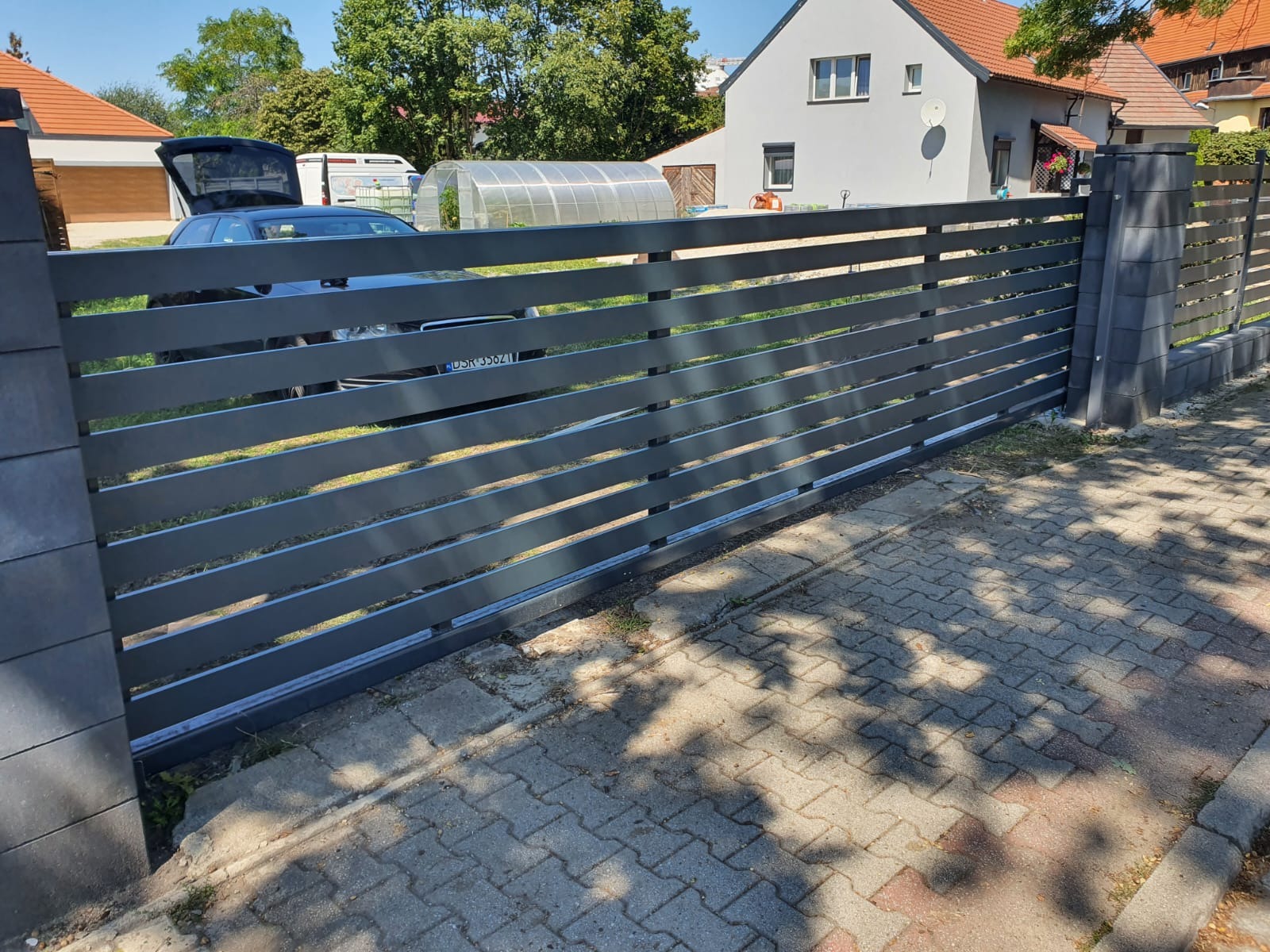 Nowoczesna przesuwna brama wjazdowa z poziomych profili, widok z zewnątrz na tle domu z ogrodem i samochodem z otwartym bagażnikiem.