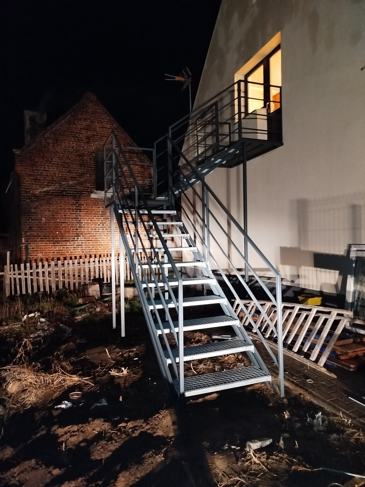 Zewnętrzne schody metalowe prowadzące do okna na piętrze budynku, widoczne przy słabym oświetleniu nocnym, z ceglanym budynkiem w tle.