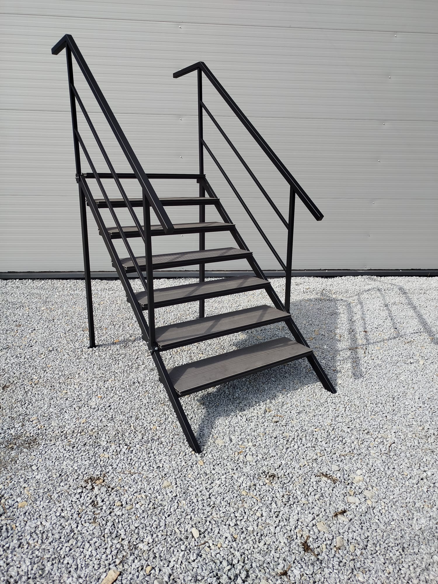 Zewnętrzne schody metalowe z czarną ramą i szarymi stopniami, ustawione na żwirowym podłożu przed szarą ścianą. Widoczne cienie schodów.
