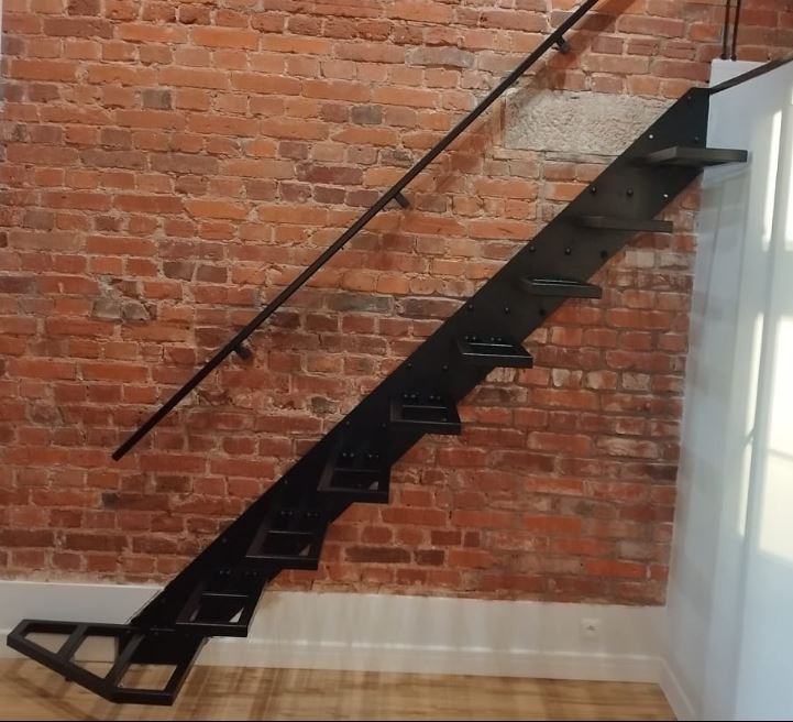 Czarne, ażurowe schody z metalu, z poręczą, zamontowane przy ścianie z czerwonej cegły, oświetlone światłem dziennym wpadającym przez okno.