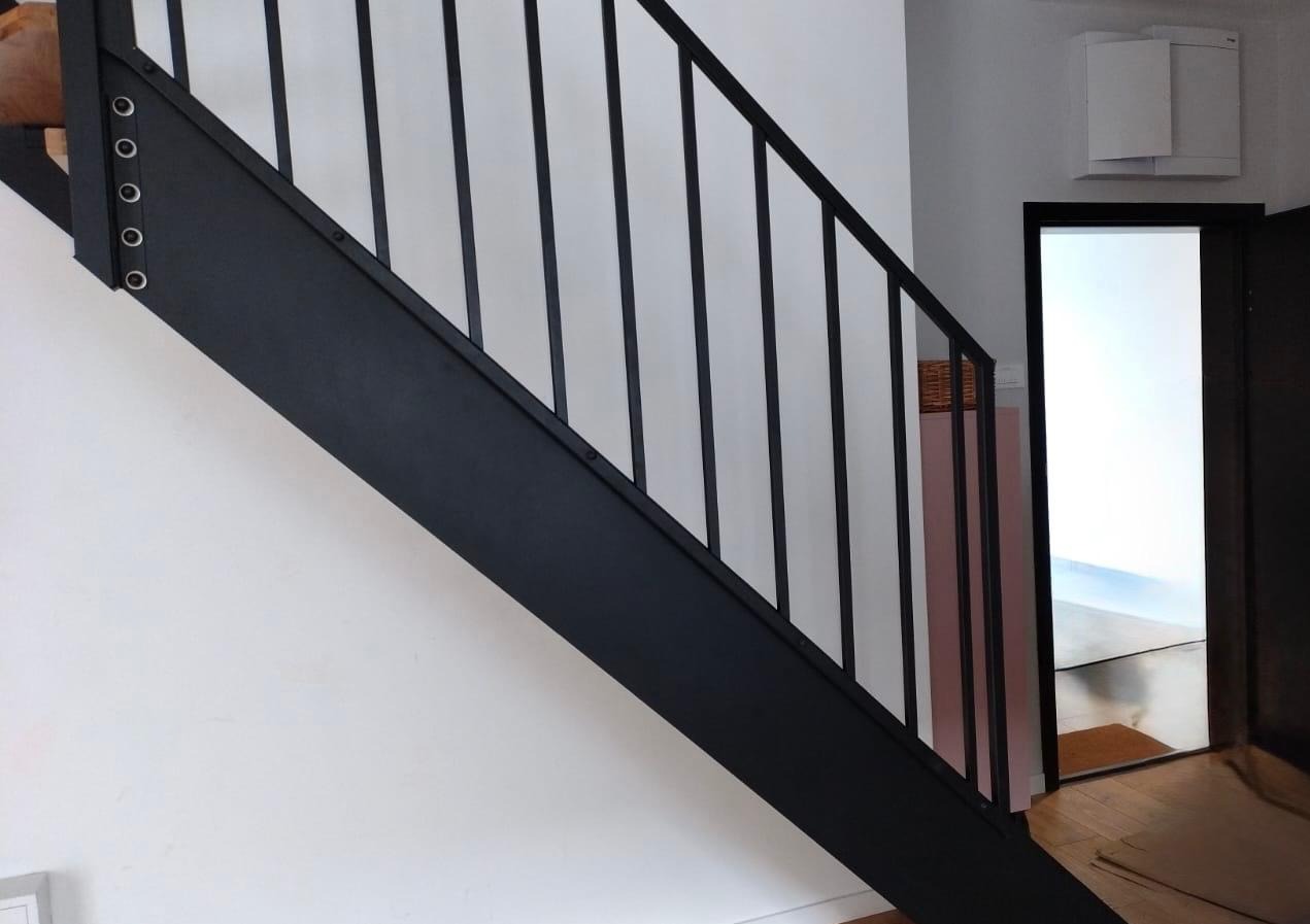 Metalowe schody z czarną balustradą z pionowymi szczeblami, mocowane na śruby, widok z dołu, białe ściany i jasne pomieszczenie w tle.