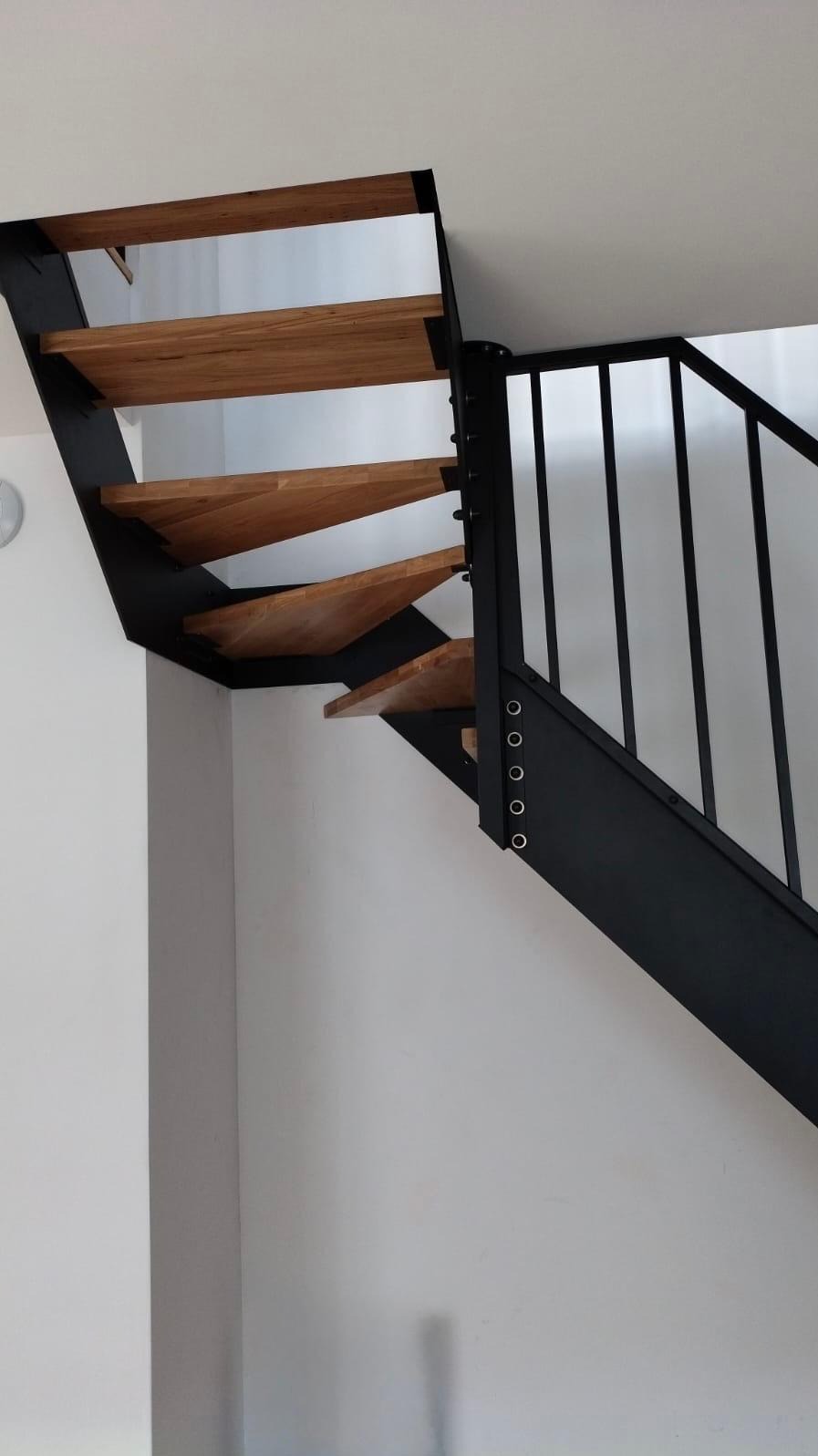 Nowoczesne schody z drewnianymi stopniami i czarną metalową konstrukcją, przymocowane do białej ściany za pomocą widocznych śrub.