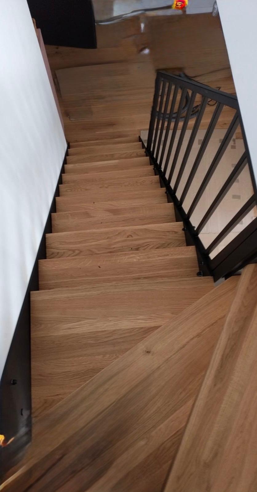 Drewniane schody z czarną metalową balustradą z pionowymi szczeblami, widoczne z góry, z fragmentami białej ściany i podłogi w tle.