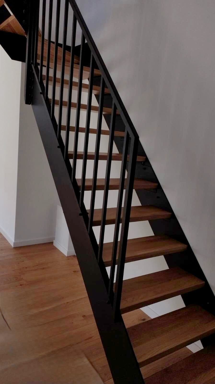 Nowoczesne schody wewnętrzne z drewnianymi stopniami i czarną, metalową balustradą, widok z dołu, jasne drewno podłogi.