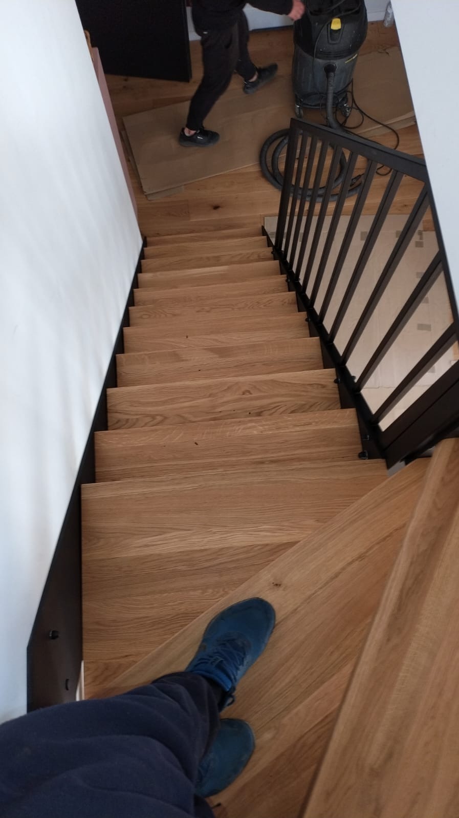 Widok z góry na drewniane schody z czarną metalową balustradą; w tle widoczna osoba z odkurzaczem przemysłowym; na pierwszym planie stopa w niebieskim bucie.